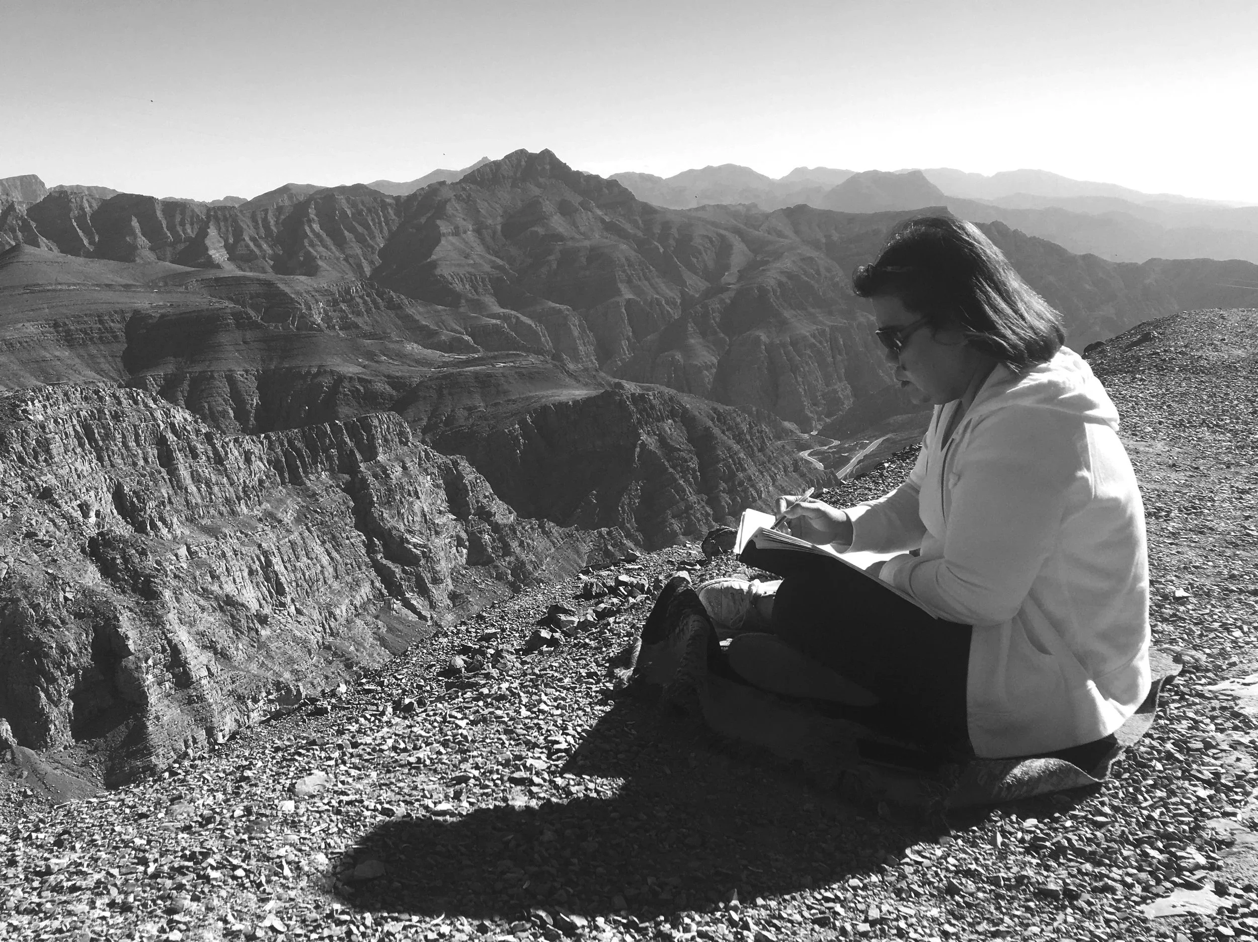 A woman sitting on a rocky ledge drawing or writing in a notebook, with a vast canyon of layered mountains in the background.