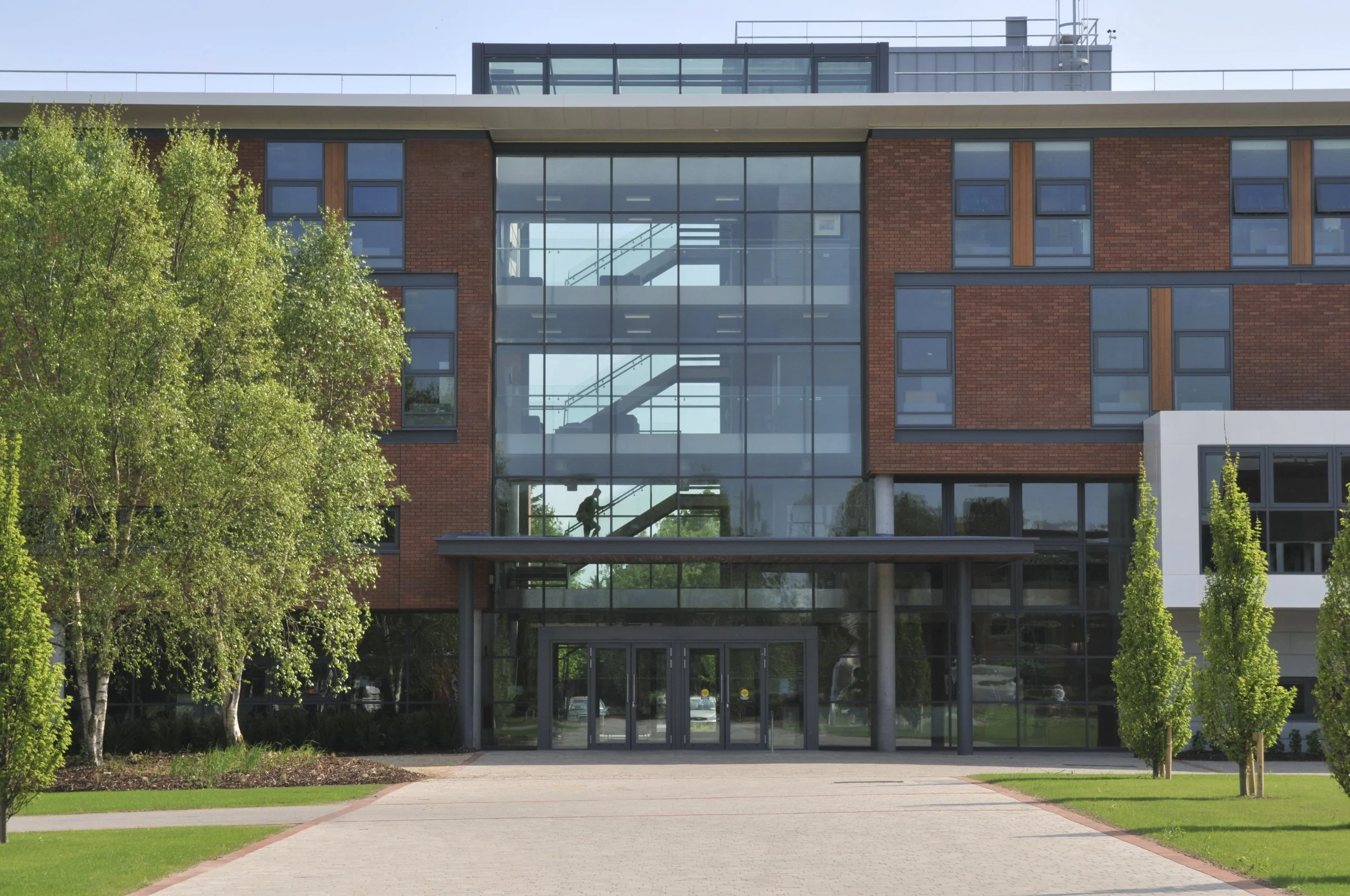 Front view of a modern building with a glass entrance, brick walls, and a staircase visible through the glass, surrounded by green trees and a landscaped walkway.