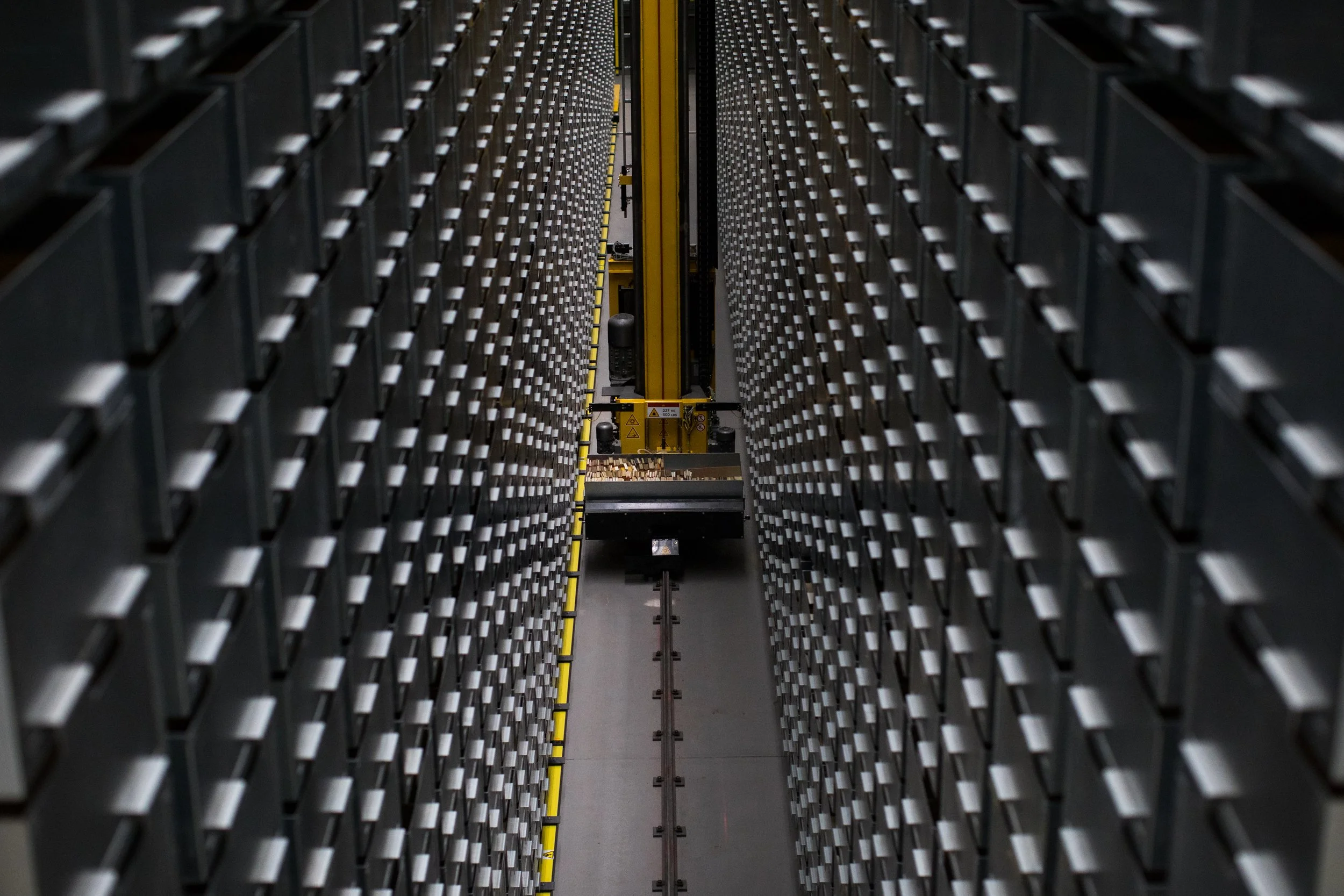 Photo of an automated storage and retrieval system with rows of trays and a yellow robotic crane in a warehouse.