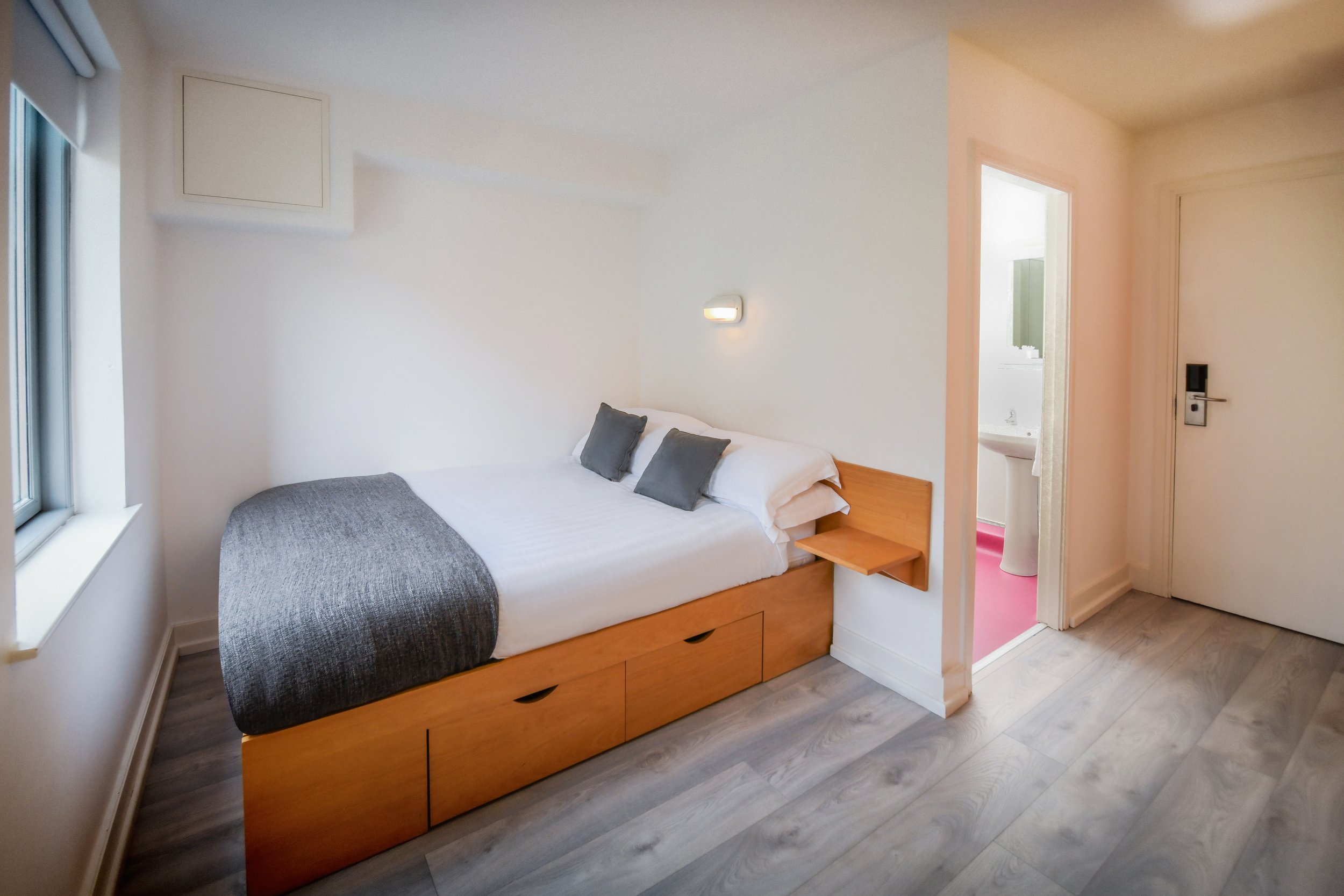 A small bedroom with a bed that has a wooden frame and drawers underneath, two gray pillows, a gray blanket, a small wooden shelf by the head of the bed, a wall-mounted light, a window to the left, an open door to a bathroom with pink flooring, a white sink, and a door to a closet on the right.