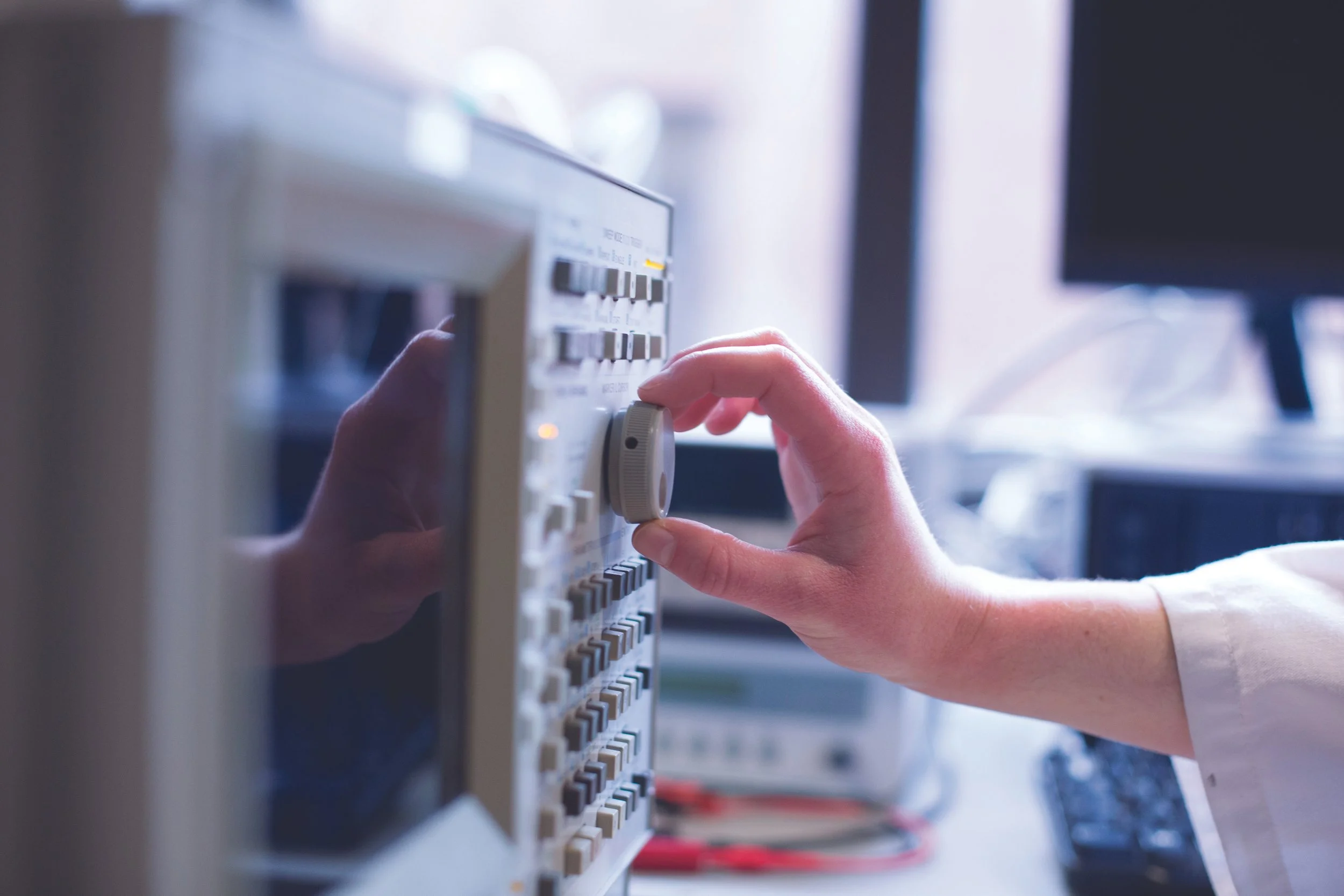 Close-up of a person's hand adjusting a control knob on an electronic testing or medical device, with other electronic equipment visible in the background.