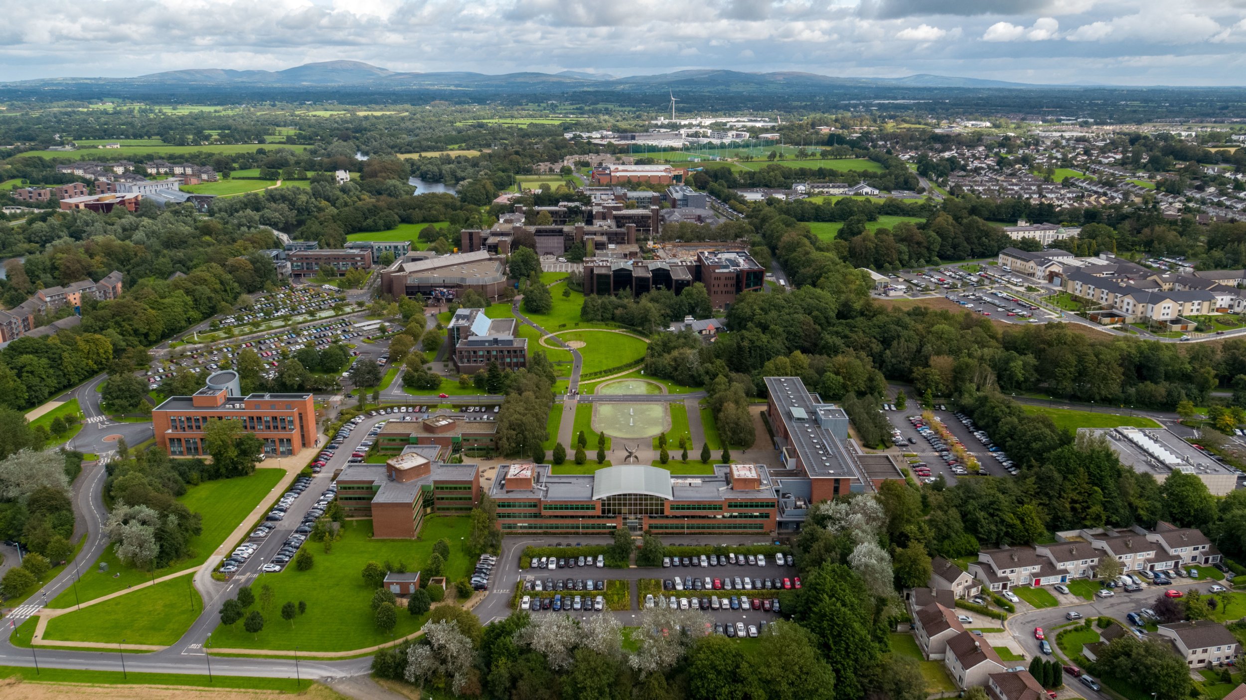 Aerial view of a university campus surrounded by green trees, parking lots, residential houses, and distant mountains under a cloudy sky.