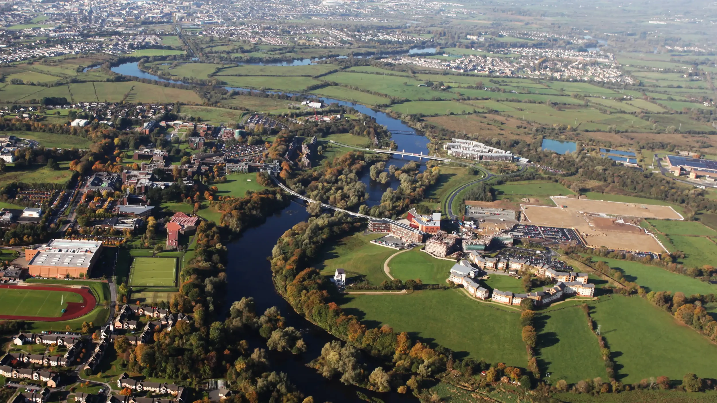 Aerial view of a city with a river flowing through green parks, buildings, and residential areas on either side.