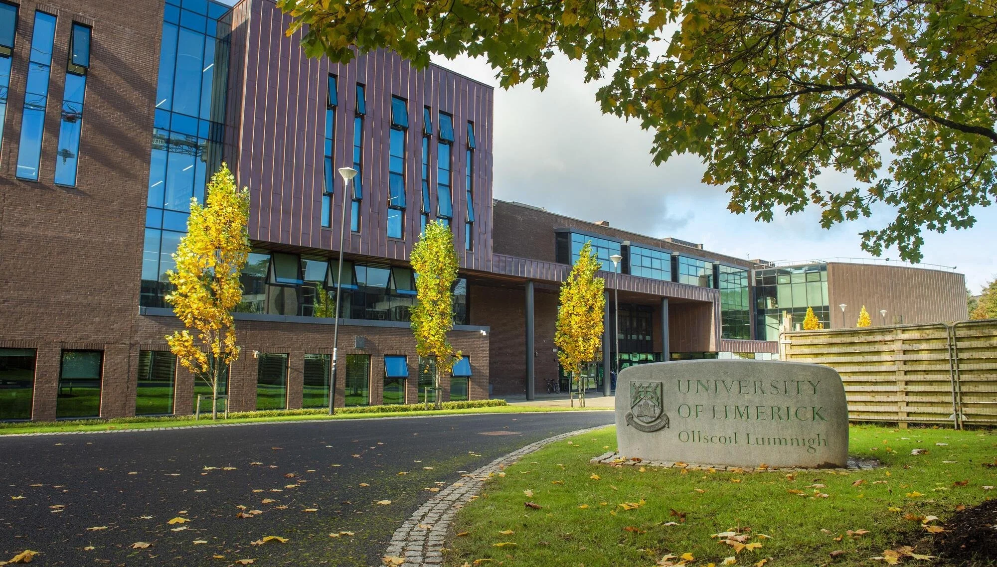 The university of limerick campus with a stone sign in the foreground, green grass, yellowing trees, and a modern brick and glass building in the background under a partly cloudy sky.