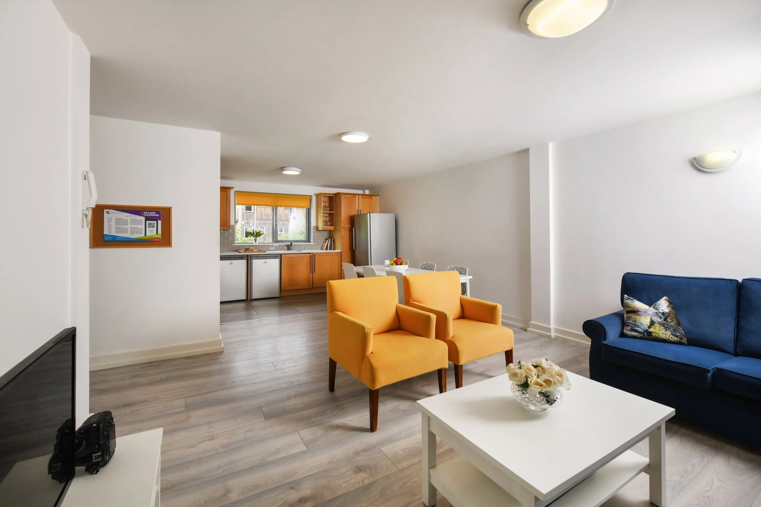 Open-concept living room and kitchen with yellow armchairs, a blue sofa, white coffee table with flowers, and a modern kitchen with wooden cabinets and stainless steel appliances.