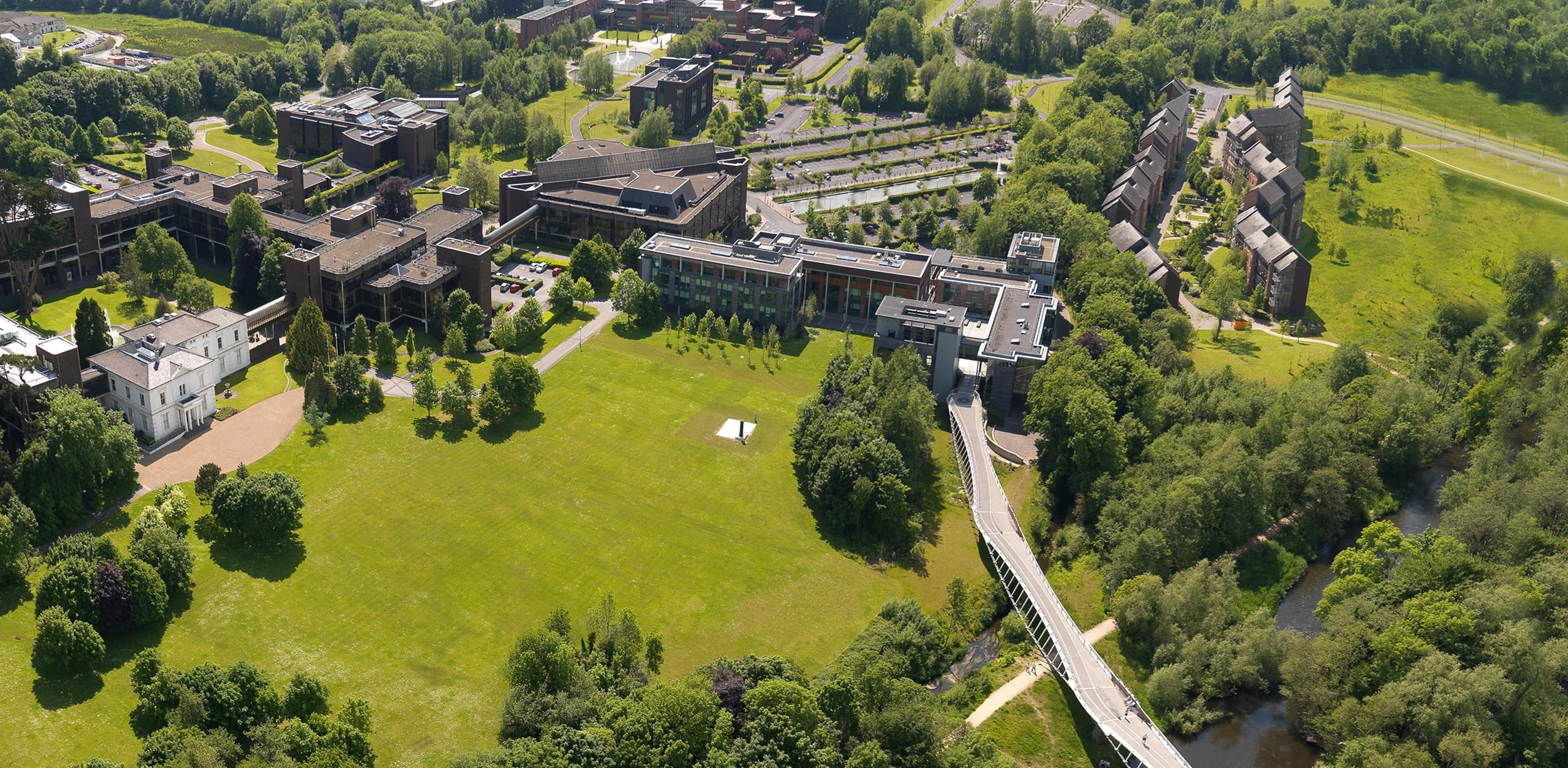 An aerial view of a college campus surrounded by greenery, including a large open grassy area, multiple buildings, a bridge over a river, and parking lots.