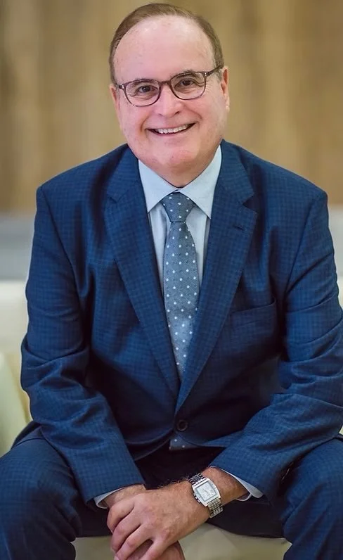 A smiling man in a blue checked suit with a light blue shirt and a polka dot tie, sitting indoors with a blurred wood background.