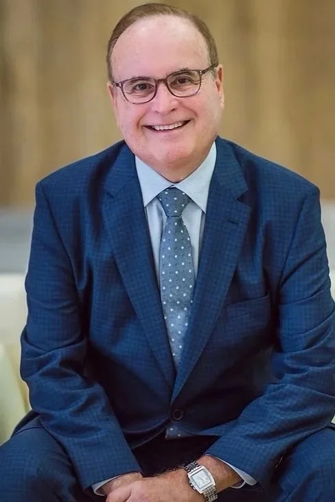 A man in a blue suit, light blue shirt, and polka dot tie, smiling, sitting indoors with a blurred background.