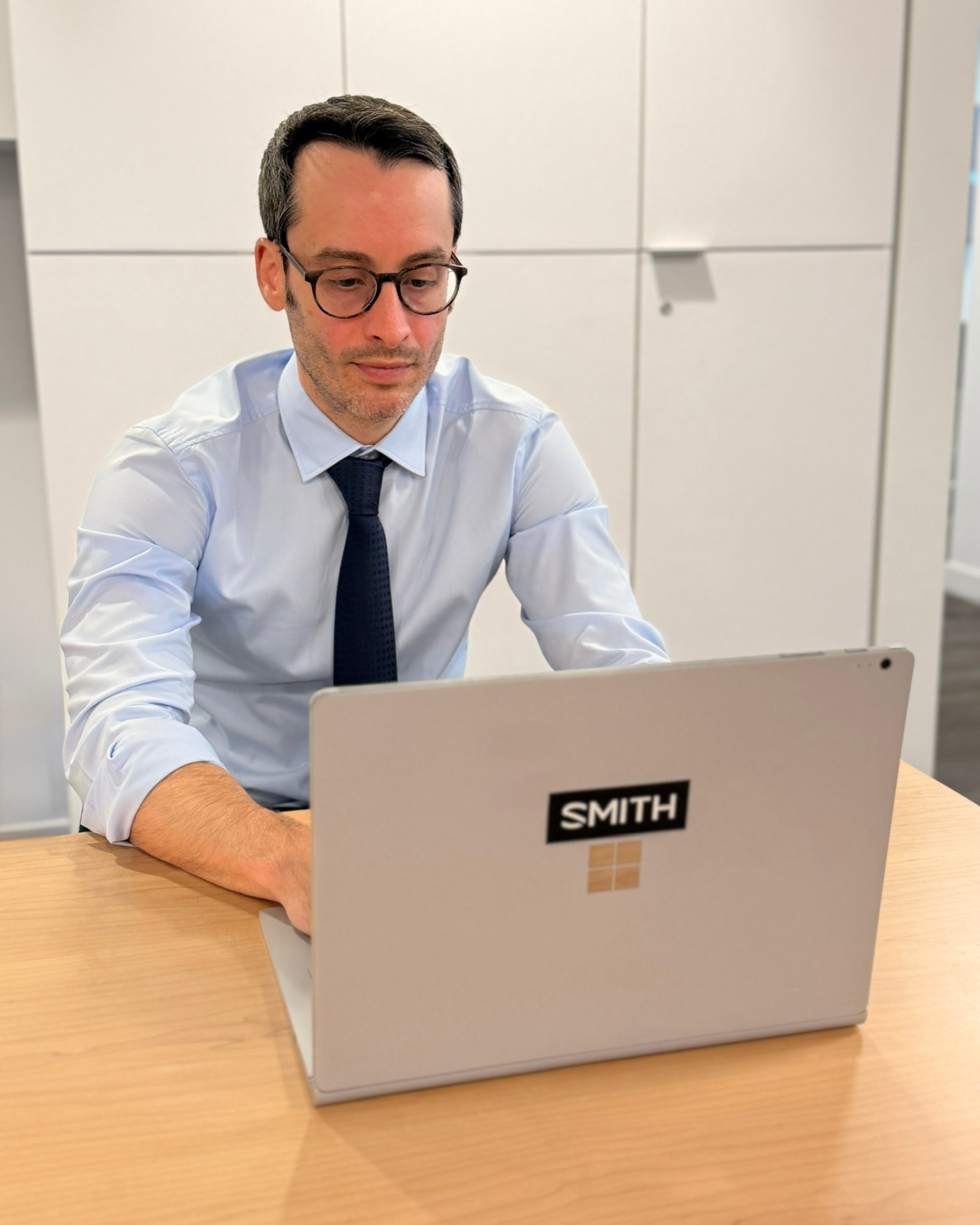 A man in a light blue dress shirt, dark blue tie, and glasses working on a silver laptop that has a black sticker with white text saying 'SMITH' and a small Windows logo. He is seated at a wooden table in an office setting with white cabinets in the background.
