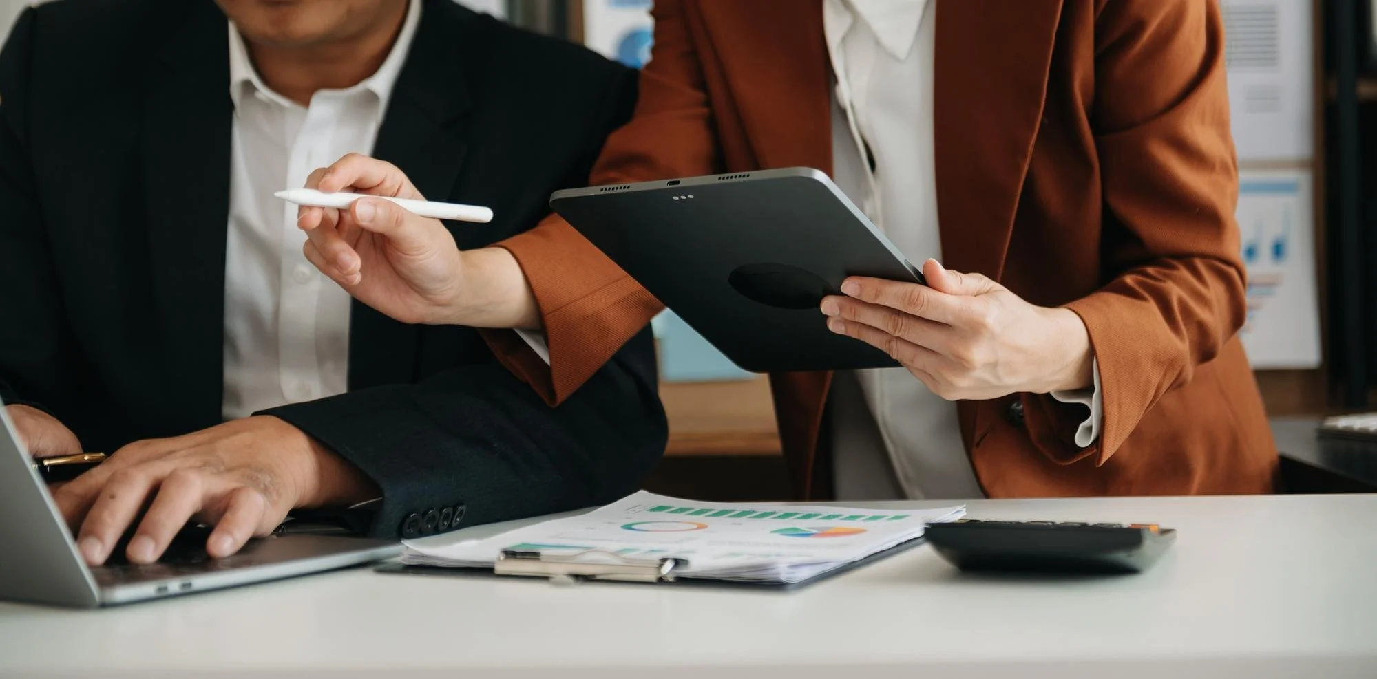 Two professionals engaged in a discussion, one person holding a tablet and the other using a stylus at a desk with documents, a laptop, and a calculator.