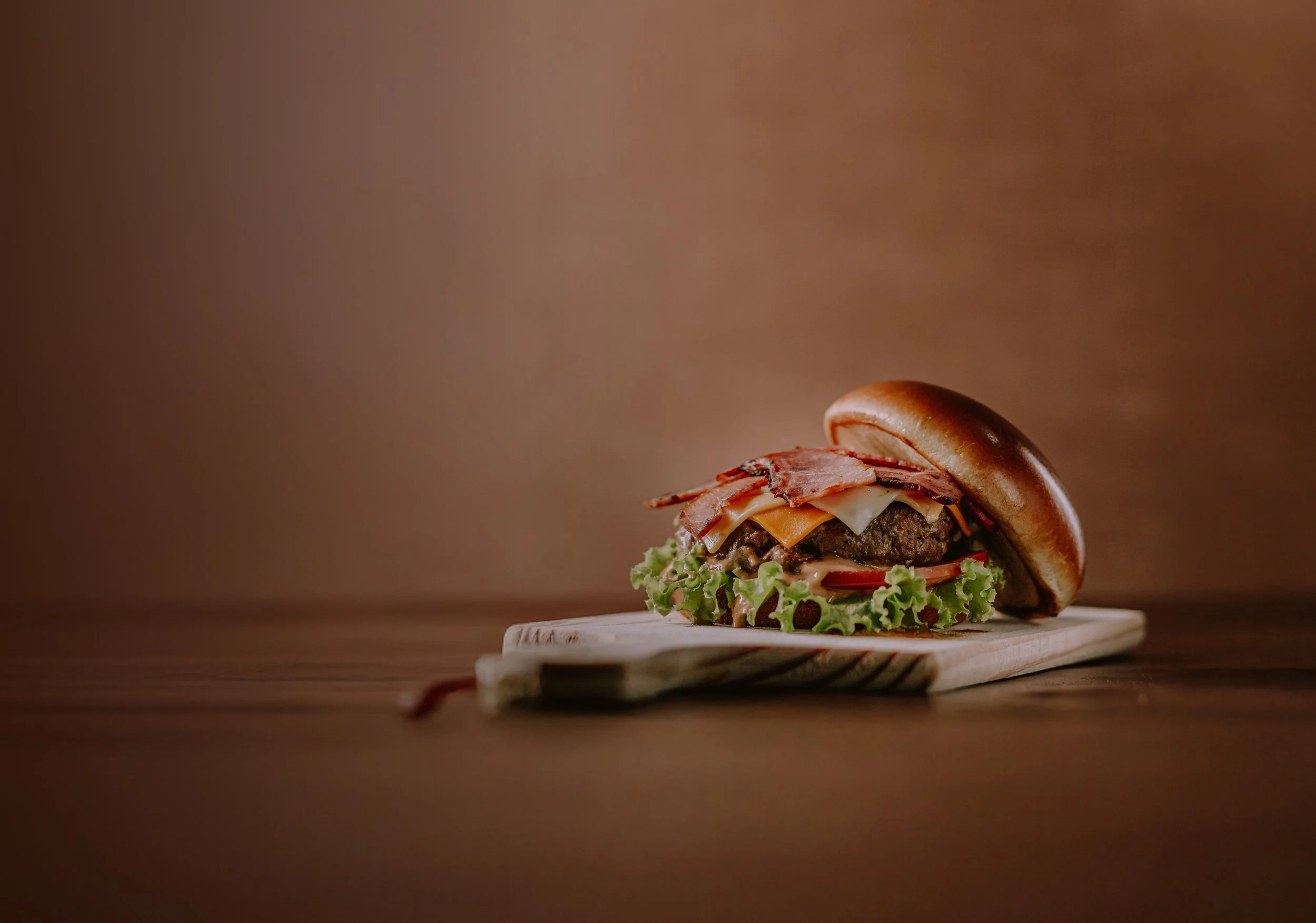 Close-up of a bacon cheeseburger on a wooden cutting board with lettuce, tomato, cheese, and crispy bacon, against a warm brown background. Fresh food in The George Stephenson pub, Newcastle.