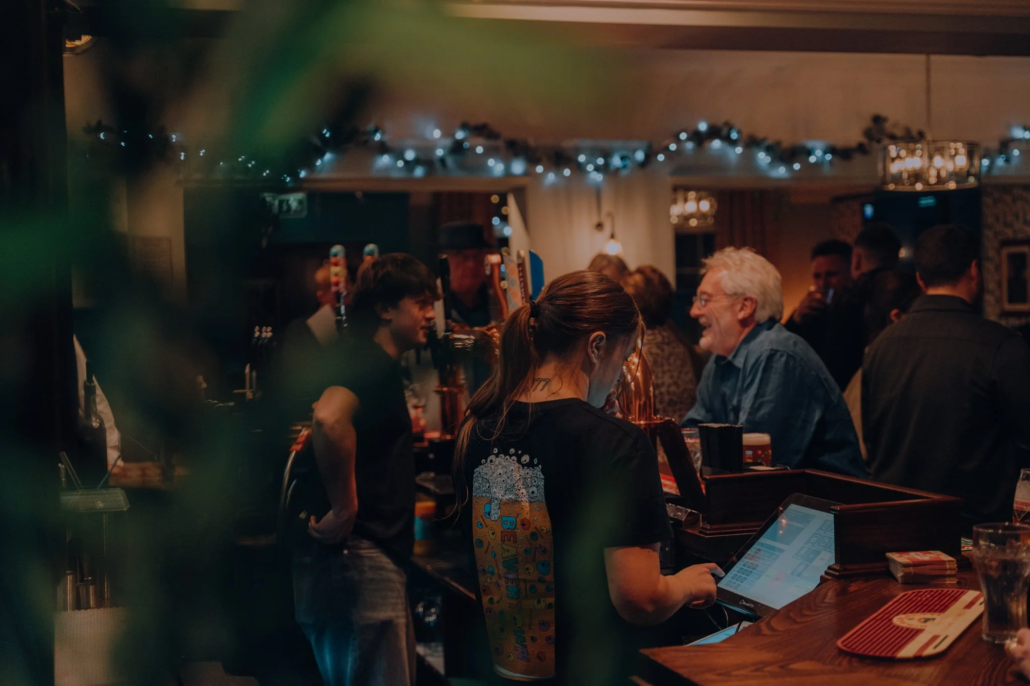 A woman working as a bartender or cashier at a pub The George Stephenson, surrounded by customers and bar tap with a wide variety of drinks and spirits. Festive Christmas string lights hanging overhead.