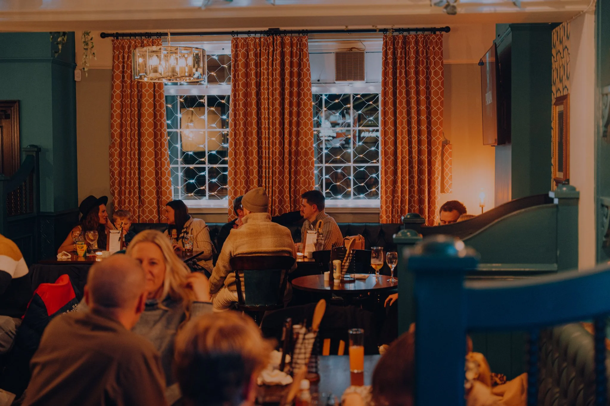 A dimly lit restaurant interior with orange patterned curtains, tables with glasses and drinks, and people dining and chatting.
