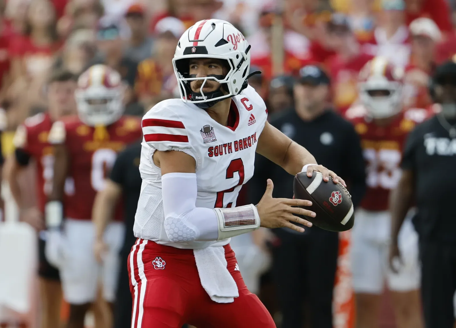 Football player in a white and red uniform with a helmet holding a football on the field, surrounded by teammates and coaches in the background.
