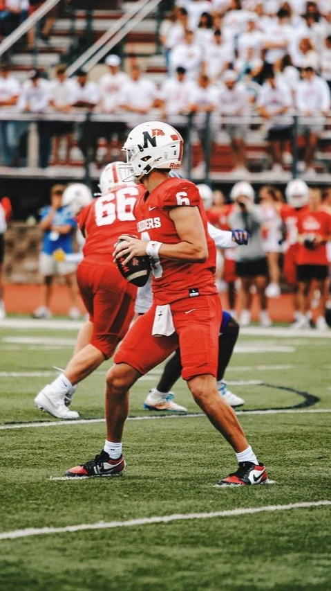 A football player in a red uniform holding a football on the field with other players and spectators in the background.