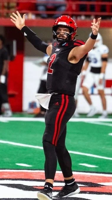 American football player celebrating with arms raised on the field, wearing a black uniform with red accents and a red helmet.