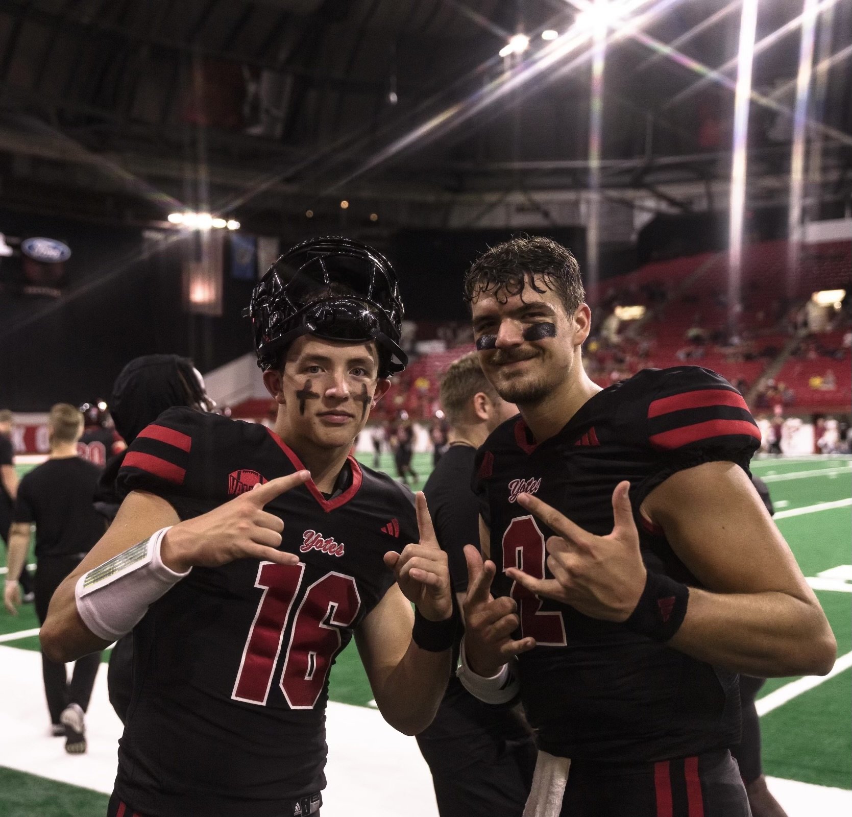 Two football players in black and red uniforms celebrating on a football field, one with a helmet and face paint, inside a stadium.