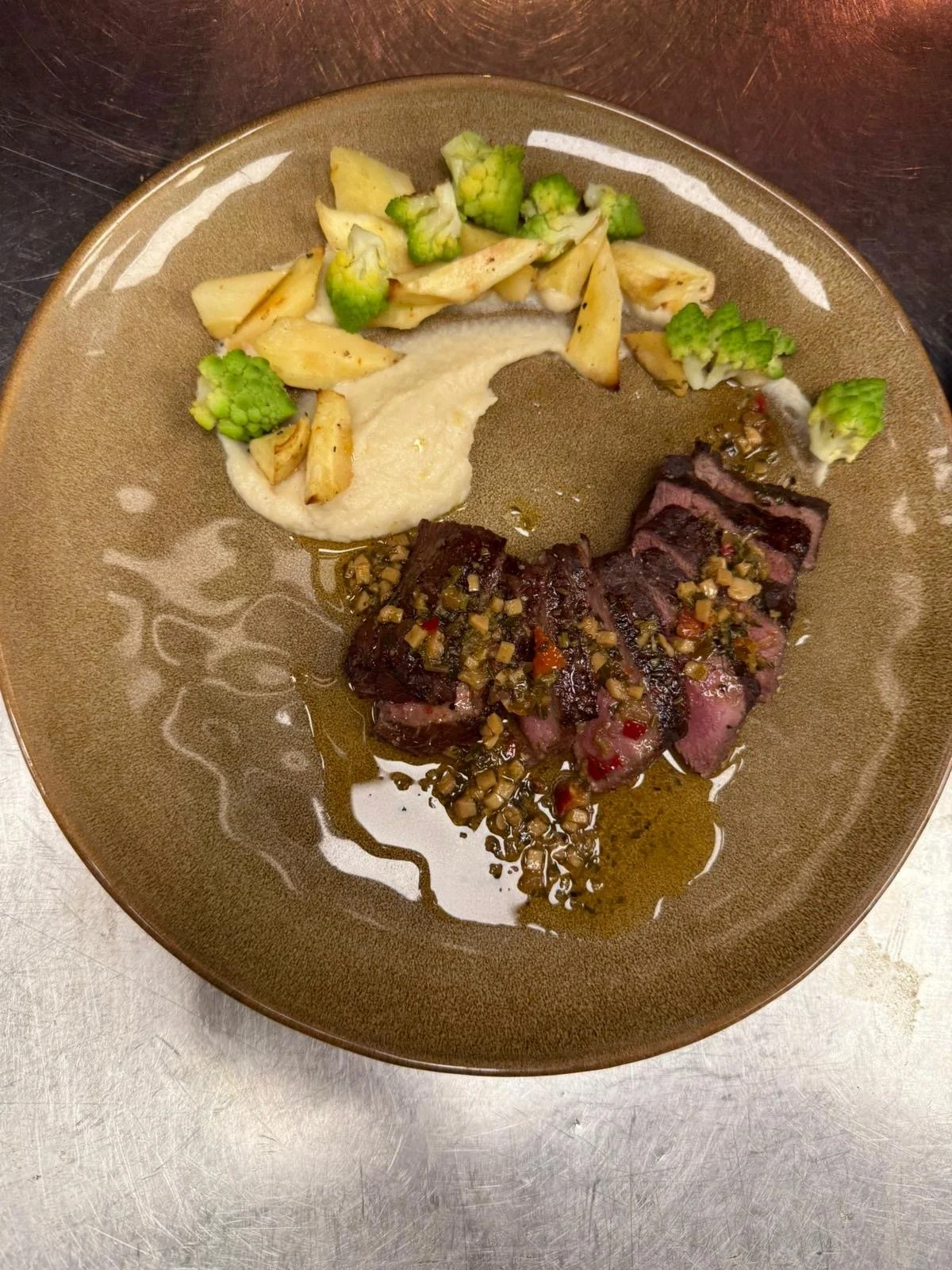 Cooked sliced beef steak with a sauce and chopped vegetables, beside broccoli and mashed potatoes on a brown plate.