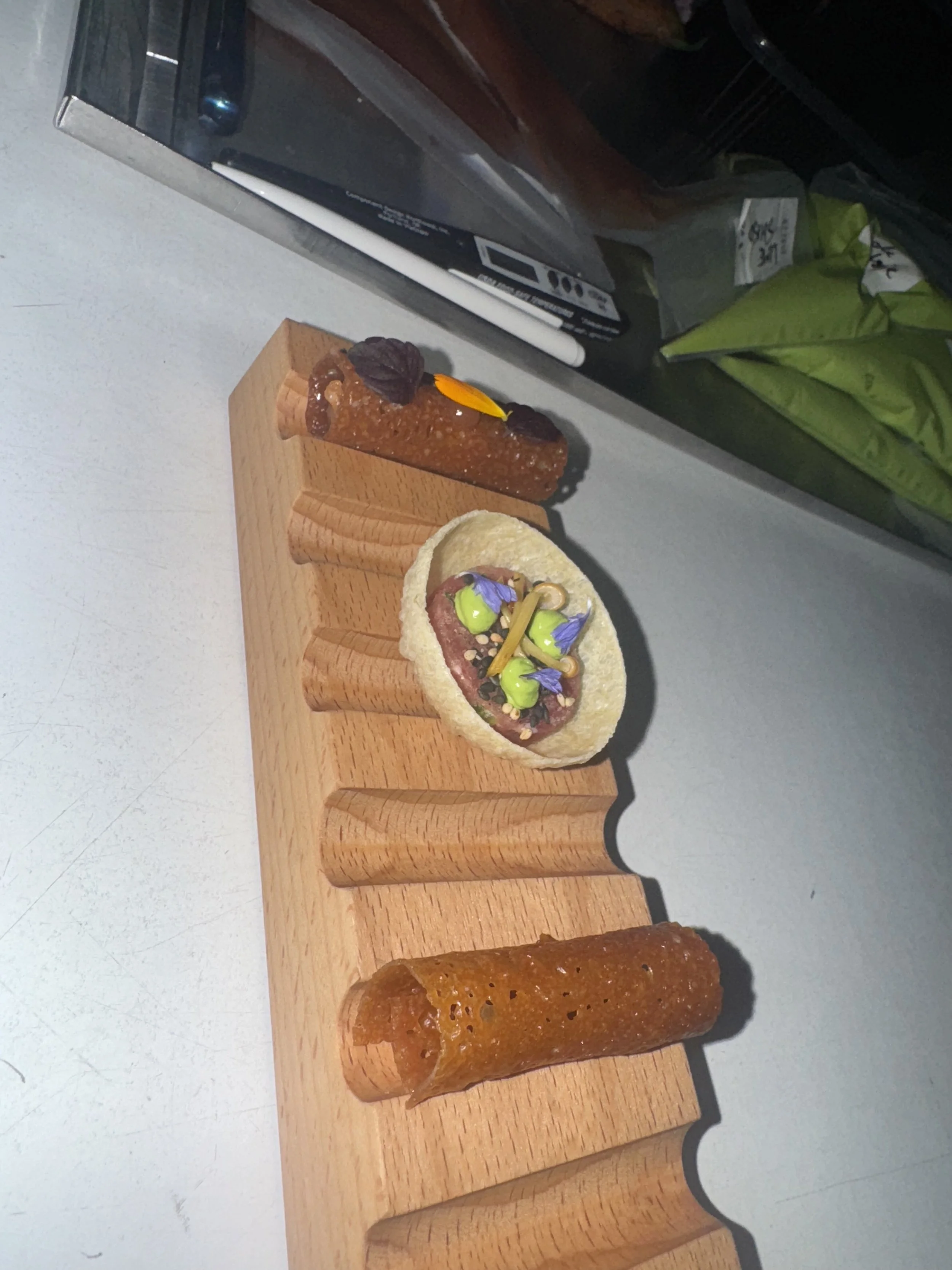 Close-up of three gourmet appetizers on a wooden serving board, with a white surface and various items in the background.