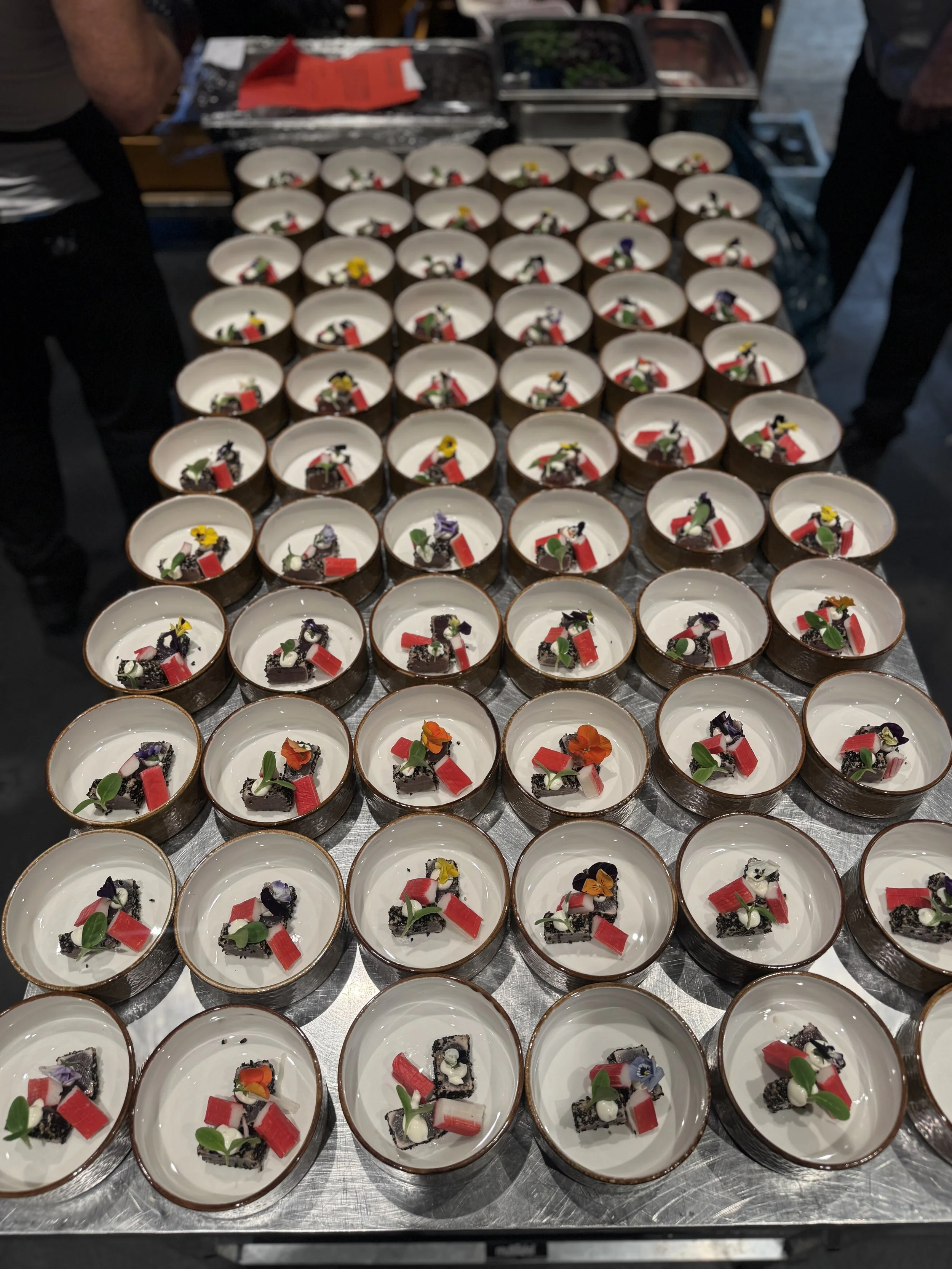 Multiple small desserts in round bowls with decorative toppings including flowers and colorful garnishes, arranged on a metallic surface.