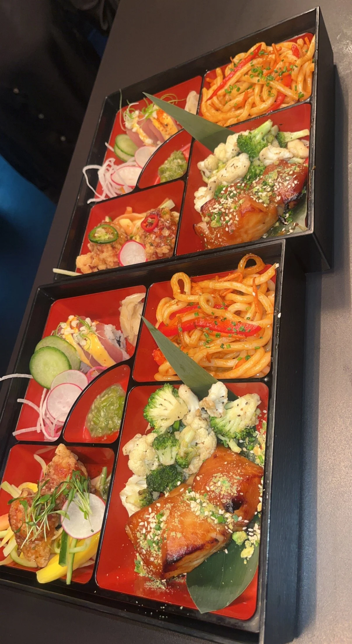 A Japanese-style bento box with various compartments containing sushi, noodles, vegetables, and fried food.
