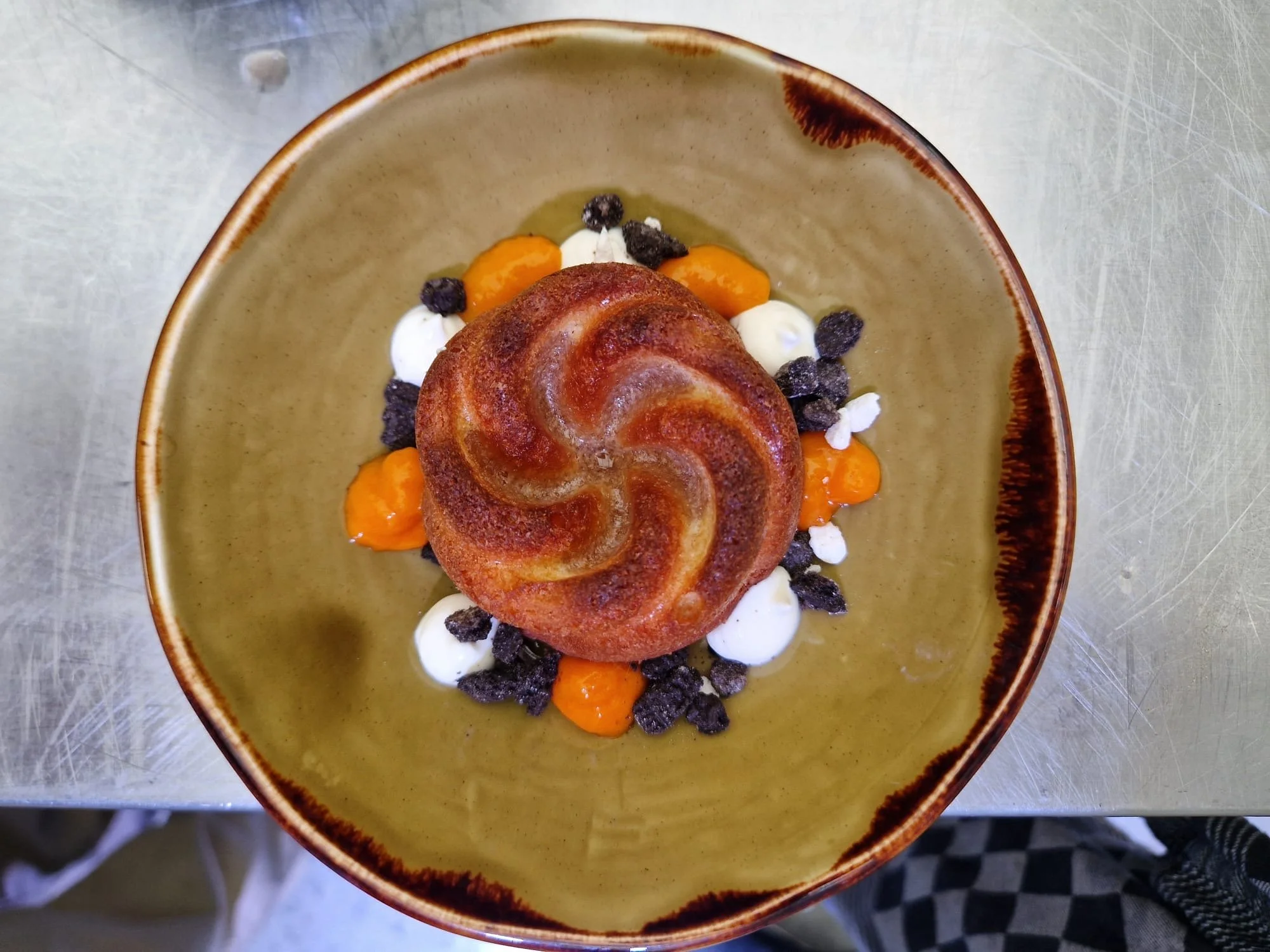 A decorative dessert featuring a swirled pastry on a bed of whipped cream, orange-colored cream, and small dark and white toppings, arranged in a rustic bowl.