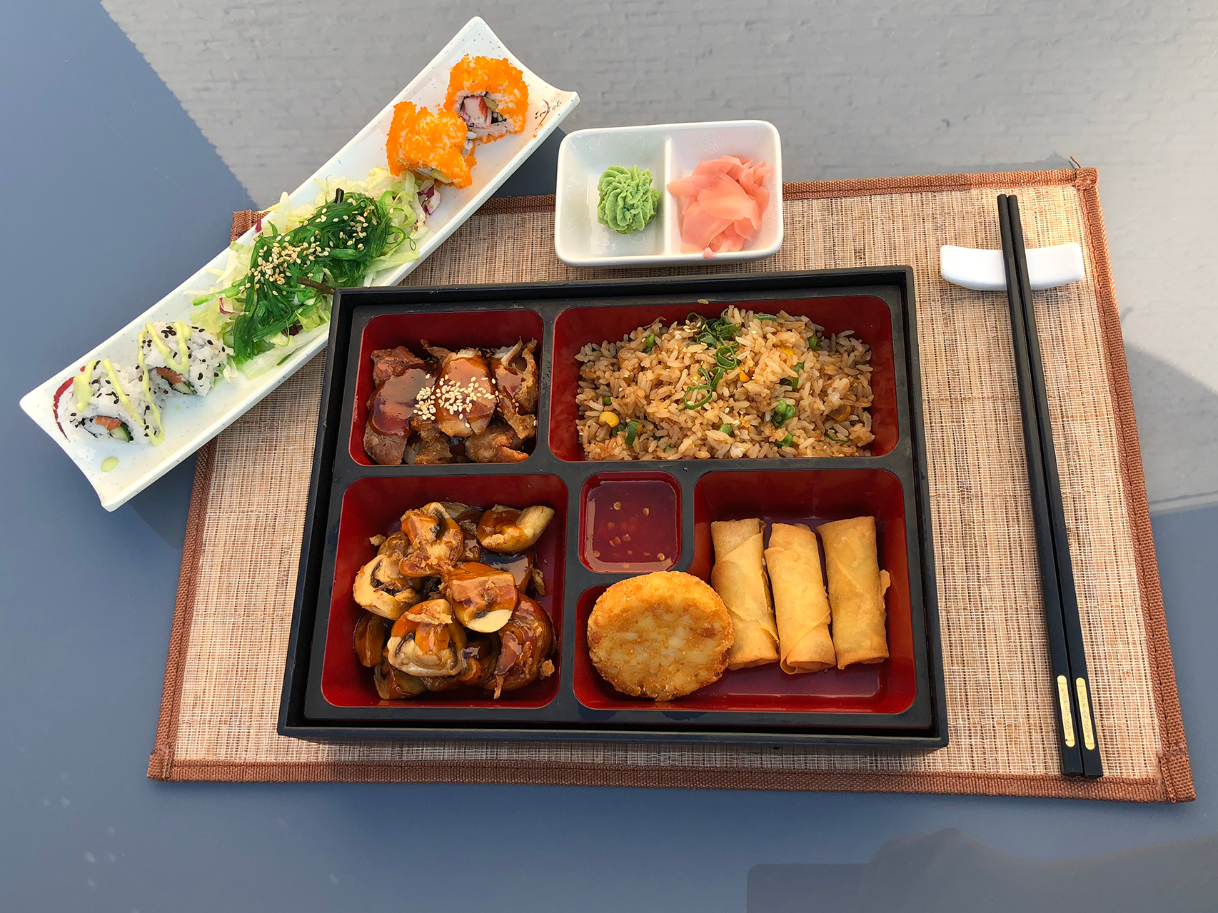 A Japanese-style lunch tray with fried rice, spring rolls, a crispy potato patty, and stir-fried vegetables, accompanied by sushi rolls with seaweed and sesame seeds, green salad with dressing, wasabi, pickled ginger, and soy sauce, with black chopsticks on a wooden placemat.