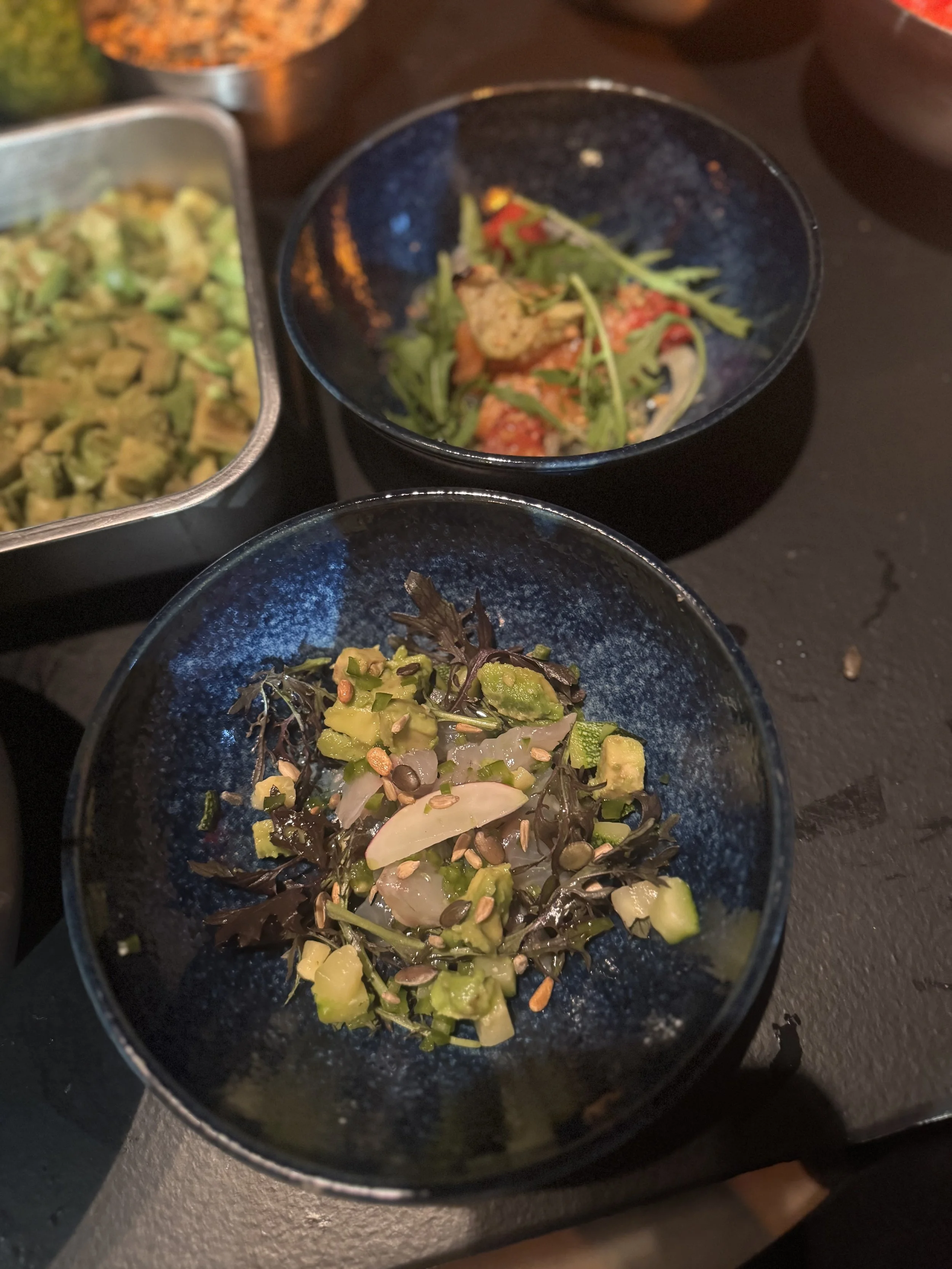 Assorted small dishes of salads with various greens, vegetables, and seeds, served in small black bowls on a dark table.