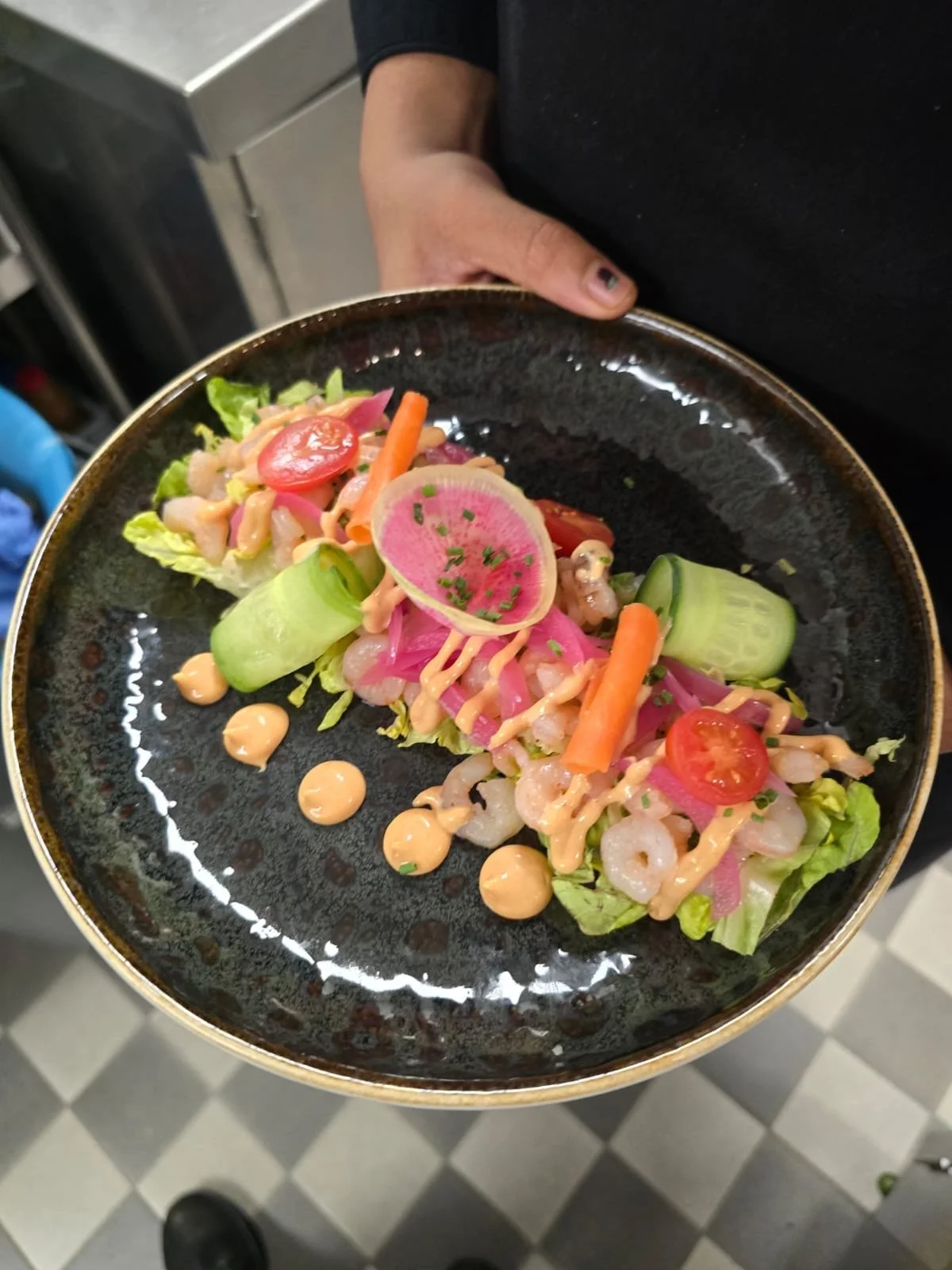 A person holding a black plate with a shrimp salad garnished with cherry tomatoes, cherry tomatoes, radish slices, cucumber rolls, shredded lettuce, and drizzled with a pink sauce.