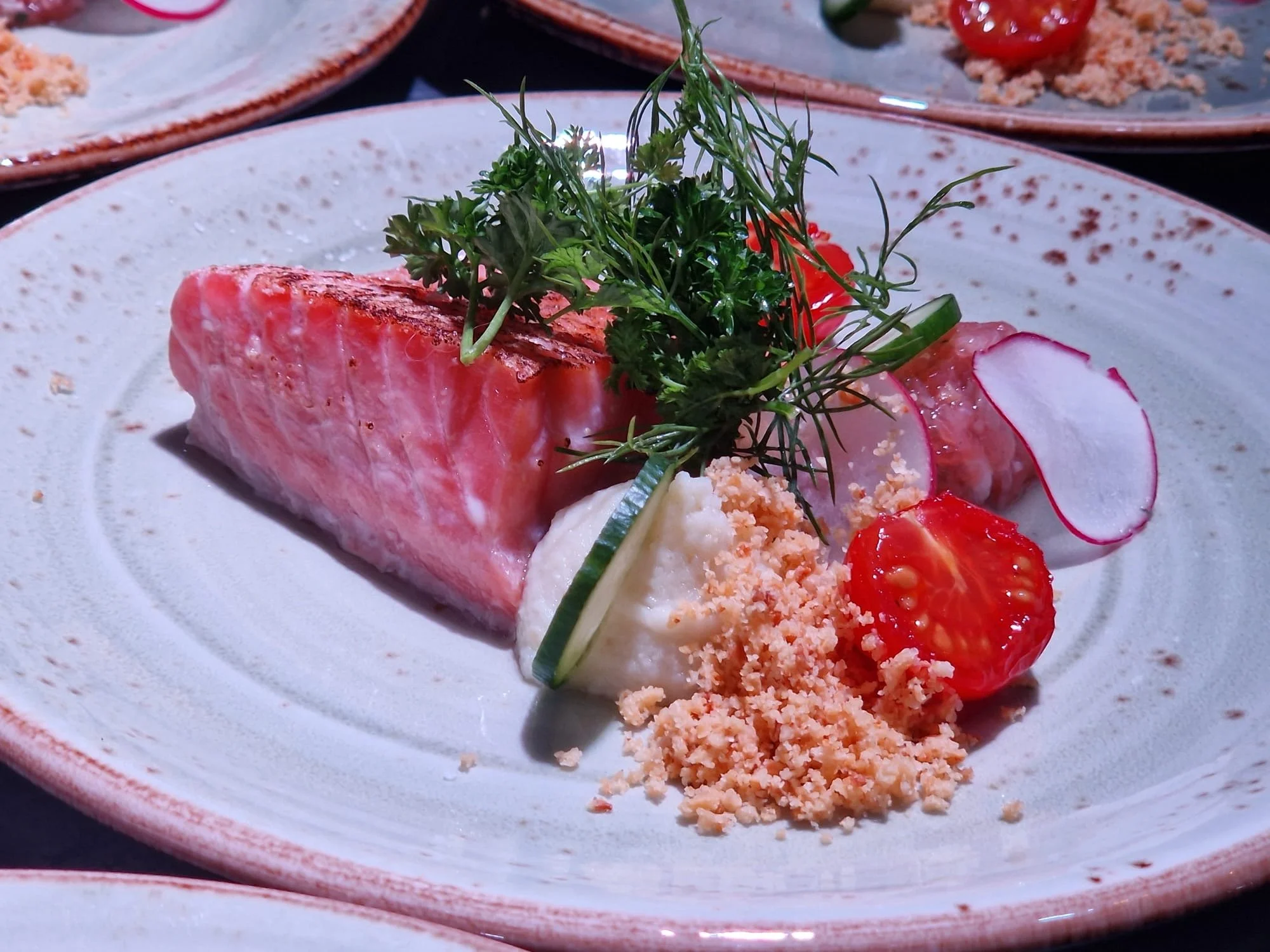 A plated dish featuring a piece of seared tuna, garnished with microgreens, radish slices, cherry tomato halves, and a side of creamy mashed potatoes topped with crumbled crackers or breadcrumb.