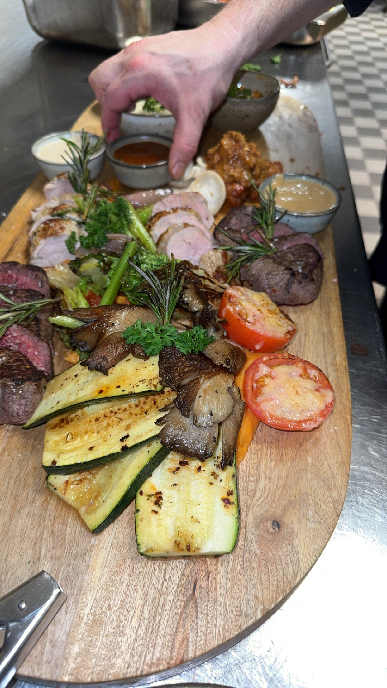 A wooden platter with grilled vegetables, sliced meats, cherry tomatoes, and garnishes, set on a metal countertop with small bowls of sauces and condiments.