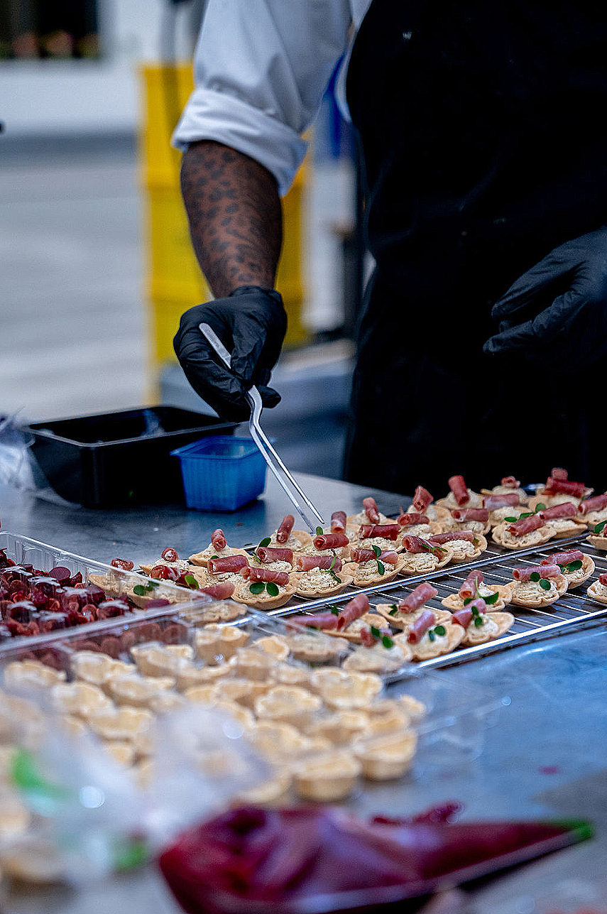 A chef or caterer preparing multiple small savory snacks with rolled ham and green garnishes on a stainless steel counter.