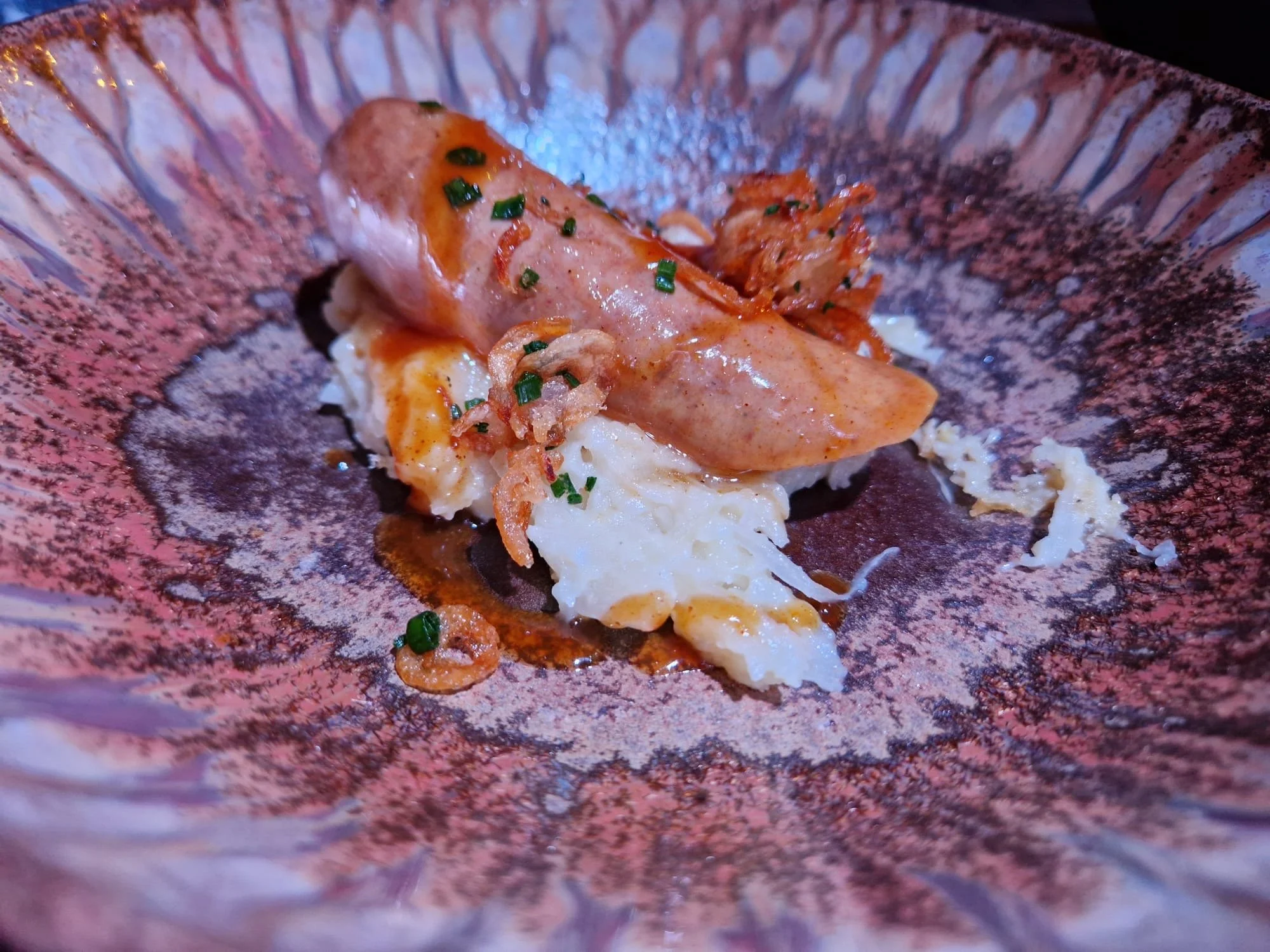 A serving of creamy mashed potatoes topped with a sausage wrapped in bacon, garnished with chopped green onions, served on a decorative pink and purple plate.