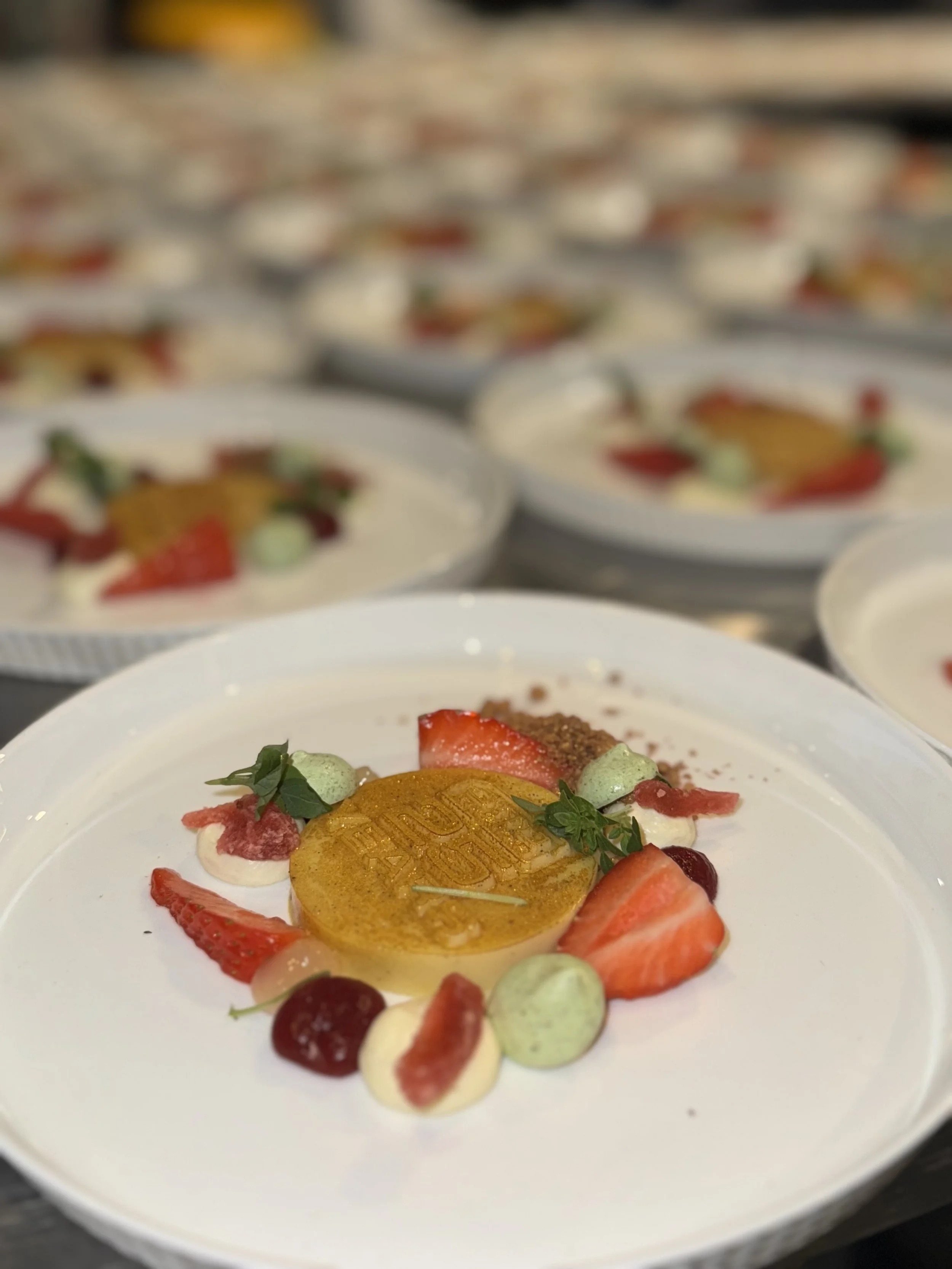A plated dessert featuring a circular yellow medallion, strawberries, cherries, dollops of cream or mousse, and garnished with small green herbs, arranged on a white plate in a restaurant setting.