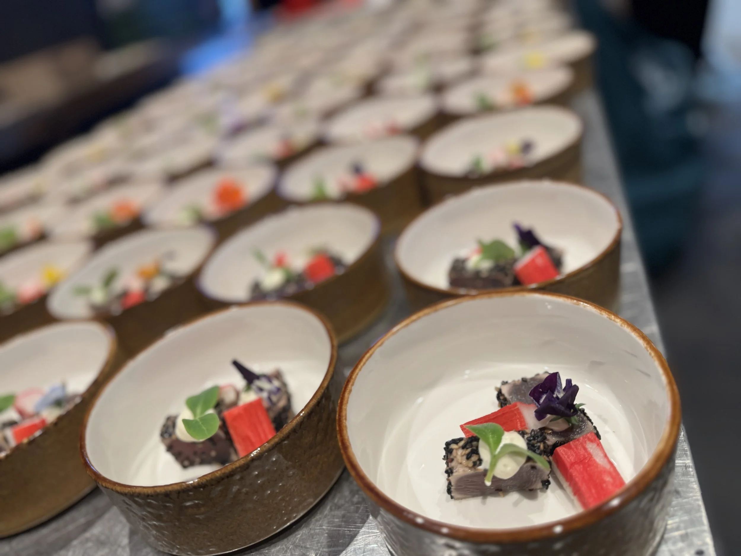Cream-colored bowls with gourmet dishes topped with herbs, red rectangular garnish, and black crumbly topping, arranged on a stainless steel surface.