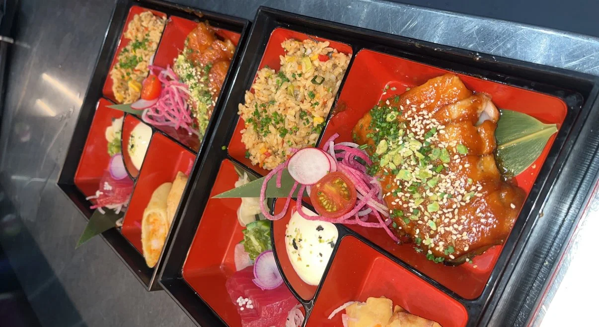 Bento boxes with rice, chicken, vegetables, and garnishes on a stainless steel surface.