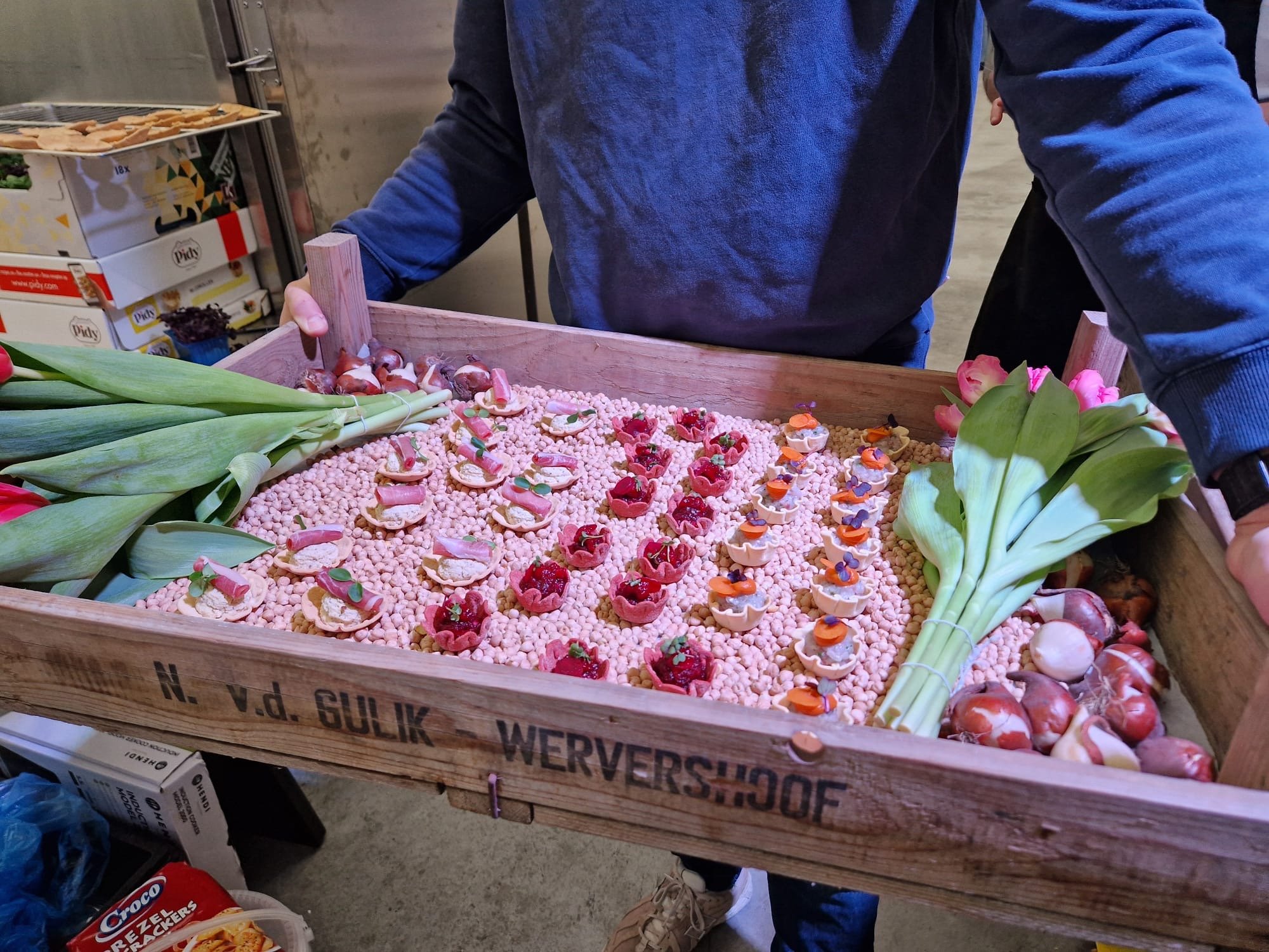 Wooden box filled with flowers, decorated treats, and pink packing balls, with person in blue shirt holding the box handle.