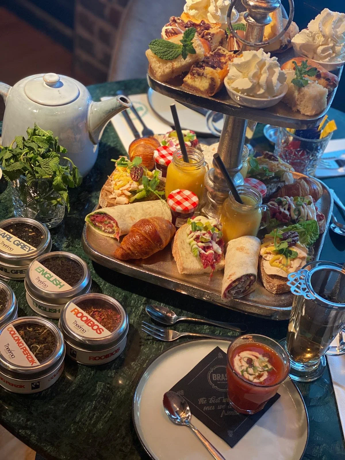 An elaborate afternoon tea table with a three-tier tray holding assorted sandwiches, pastries, and desserts, a teapot, small jars of jam, and cups of tea. There are also containers of tea leaves labeled with different flavors, and a glass of Bloody Mary with garnishes.