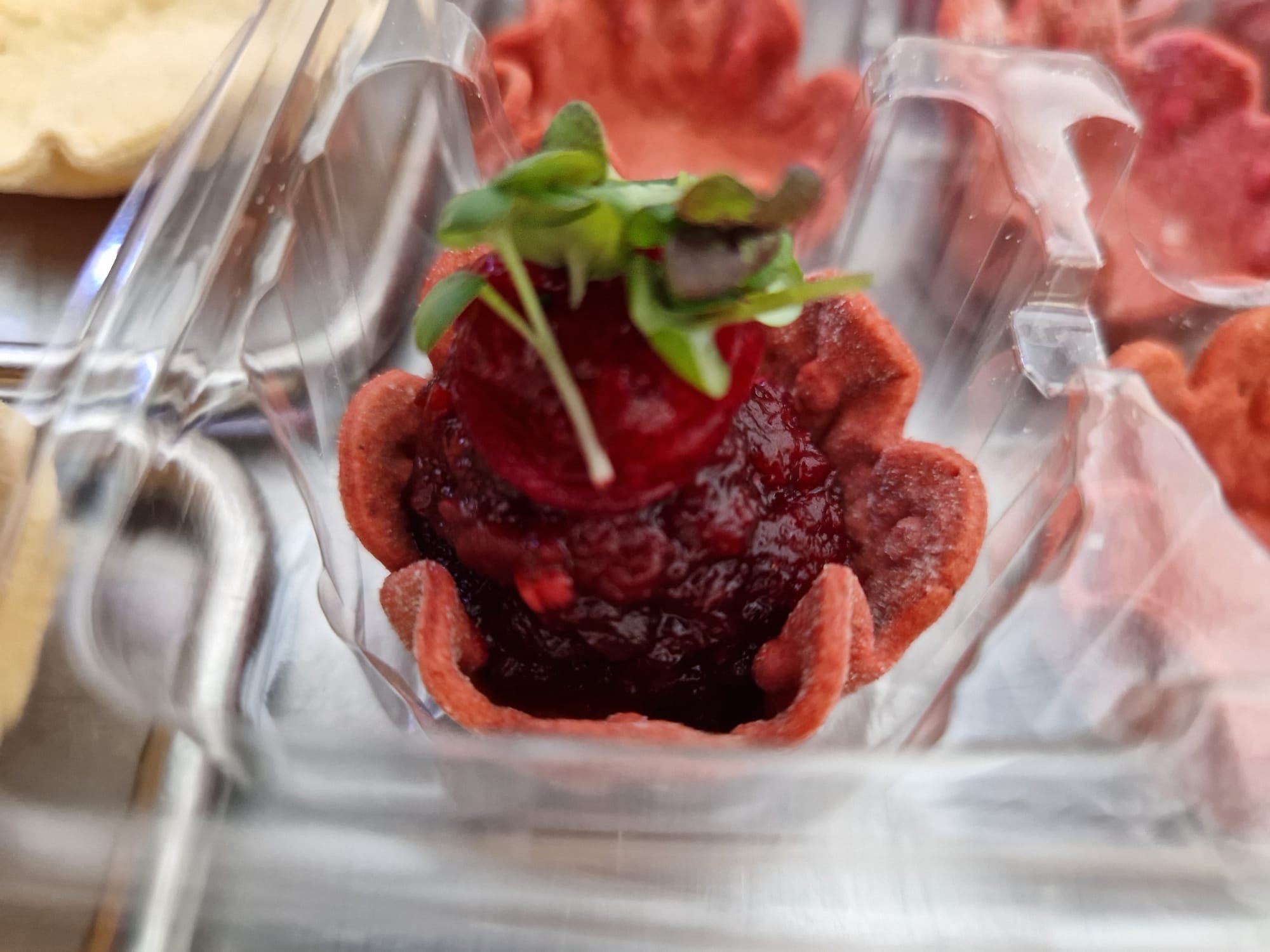 Close-up of a mini tart filled with berry jam, topped with a fresh raspberry and microgreens, in a clear plastic container.