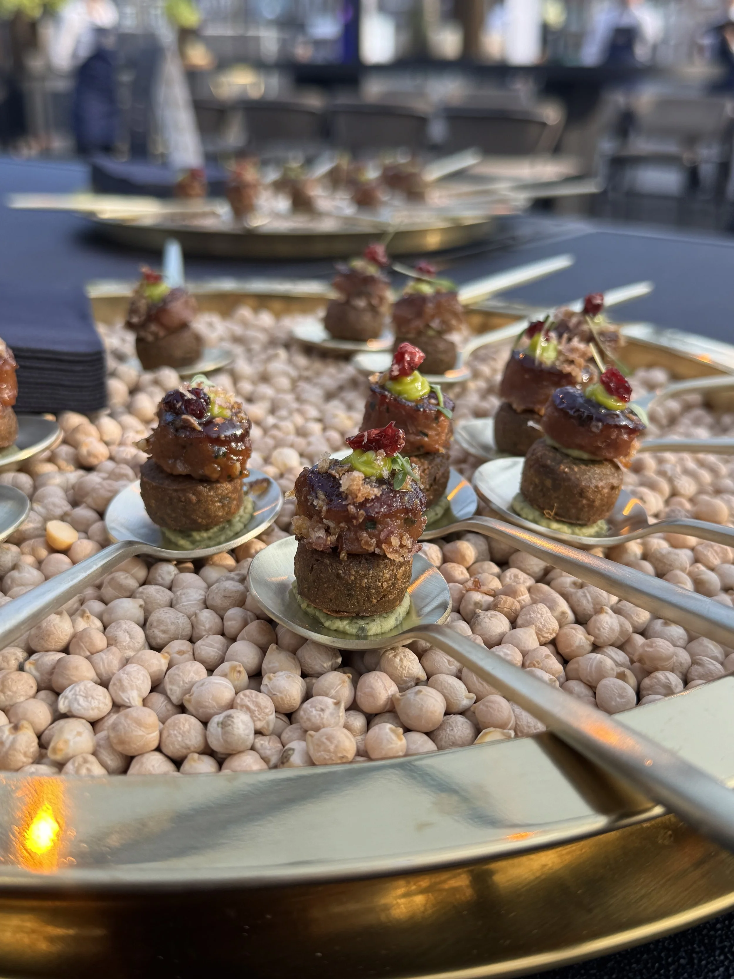 Close-up of bite-sized appetizers on spoons, arranged on a bed of white pebbles at an outdoor event, with a blurred background.