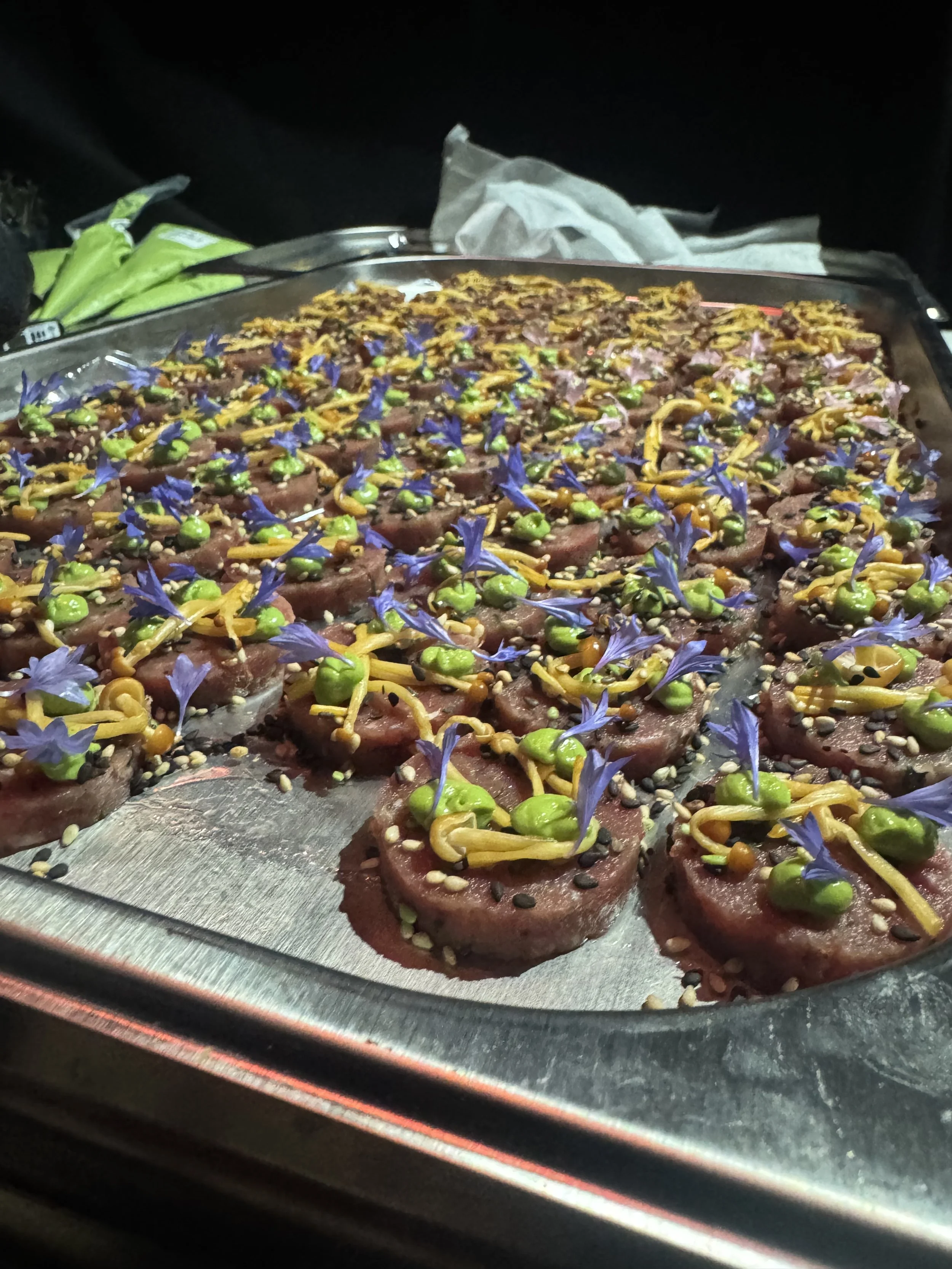 Close-up of raw beef tartare topped with edible flowers, capers, and seeds on a metal tray in a dimly lit setting.