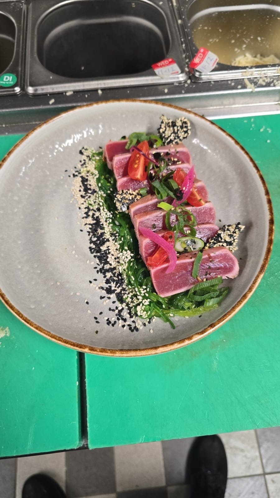 A plate of seared tuna sashimi garnished with sliced green onions, cherry tomatoes, pickled ginger, sesame seeds, and seaweed, served on a gray ceramic dish.