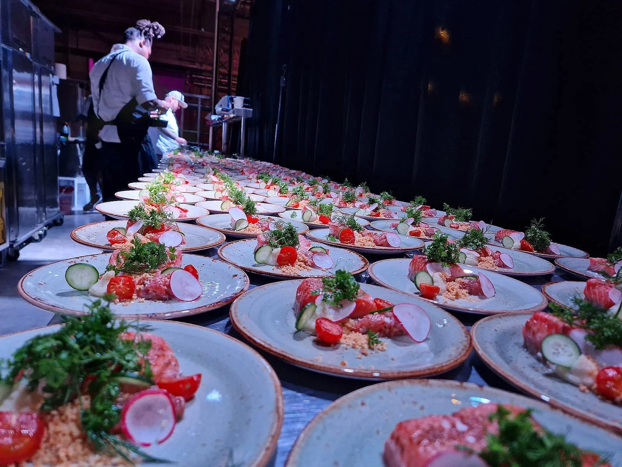 Several plates of salmon garnished with cherry tomatoes, sliced radishes, and herbs are laid out in a row on a long table, with people preparing food in the background.