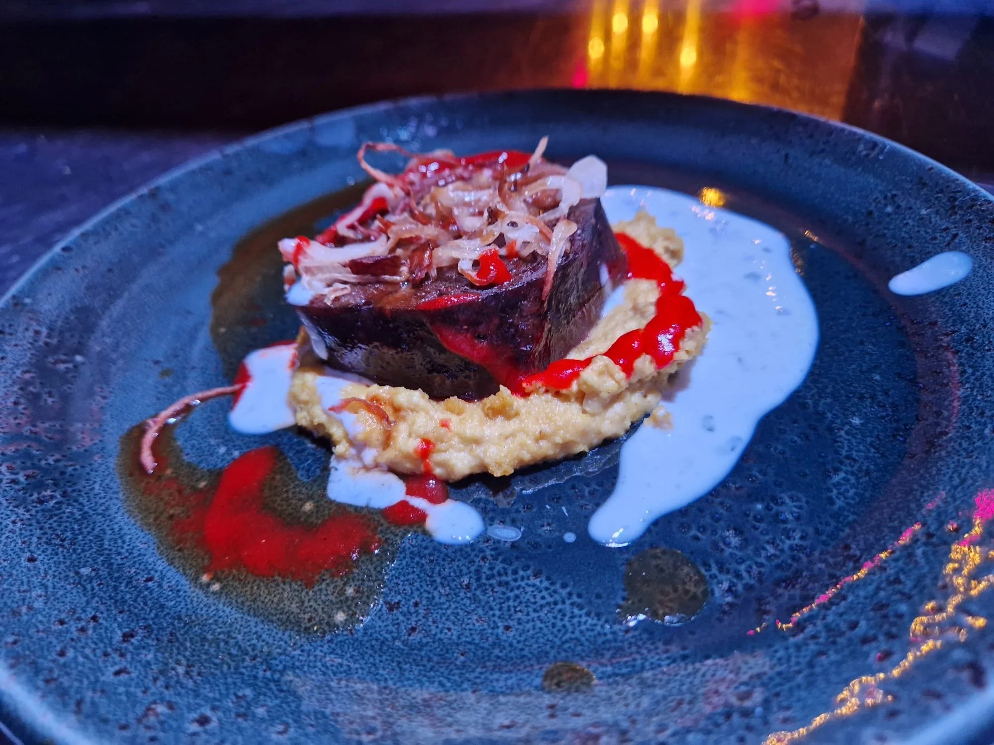 A plated appetizer featuring a square piece of meat or terrine topped with shredded vegetables, served on a bed of sauce with white and red sauces drizzled on the black plate.