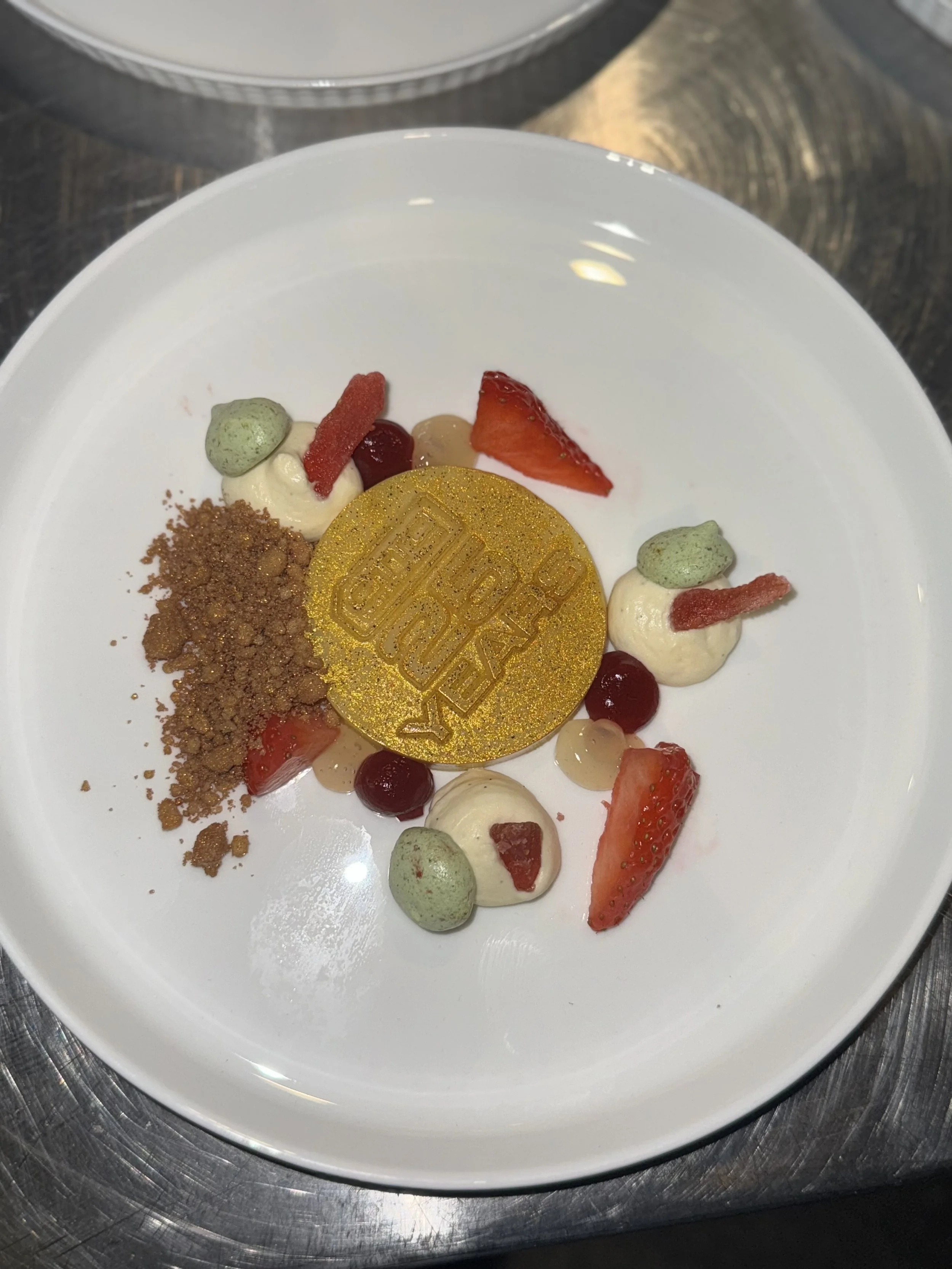A dessert plate with strawberries, red and black berries, small dollops of cream, green powder, brown crumble, and a gold disc with the word 'PERFECT' embossed on it.