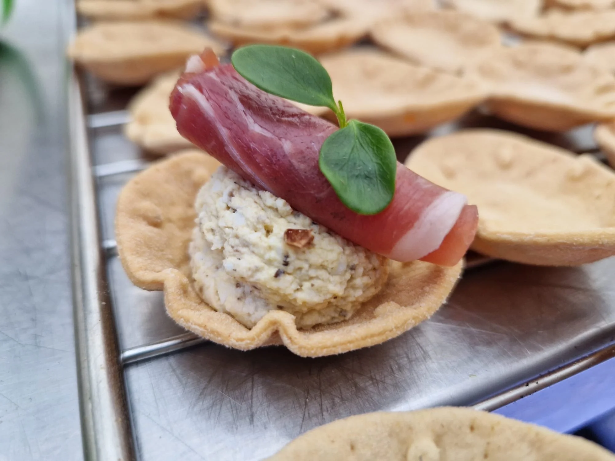 A close-up of a canapé with a tart shell filled with cheese, topped with a rolled slice of prosciutto and a small green leaf, on a metal tray.
