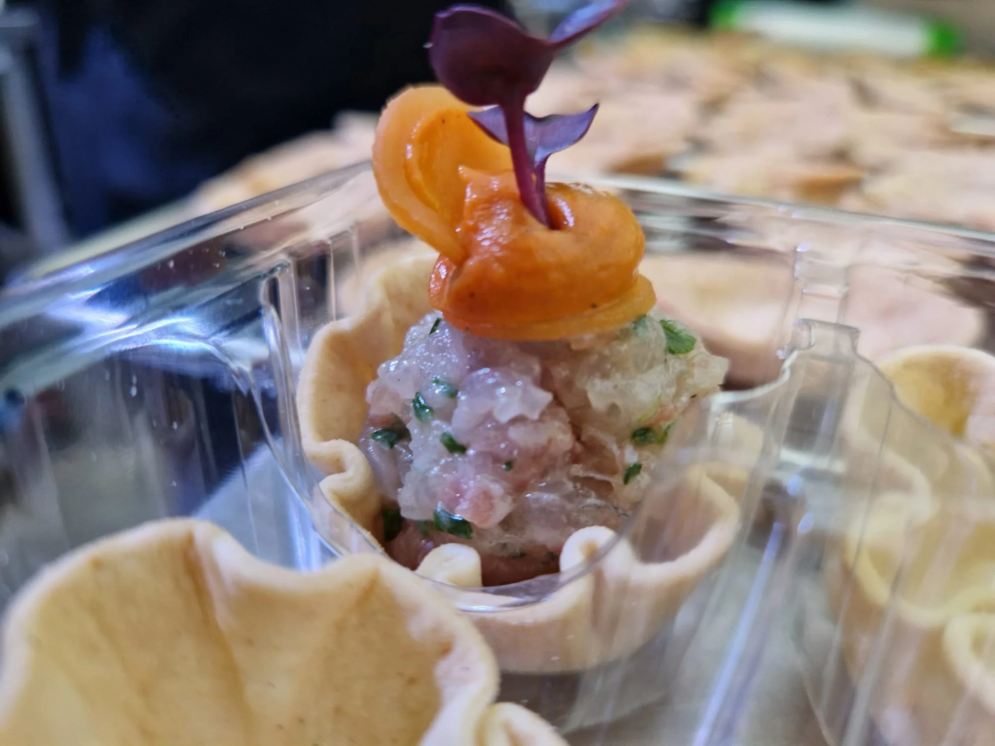 Close-up of a seafood appetizer in a plastic cup with shredded ice, topped with a slice of orange and a purple edible flower, surrounded by small tart shells.