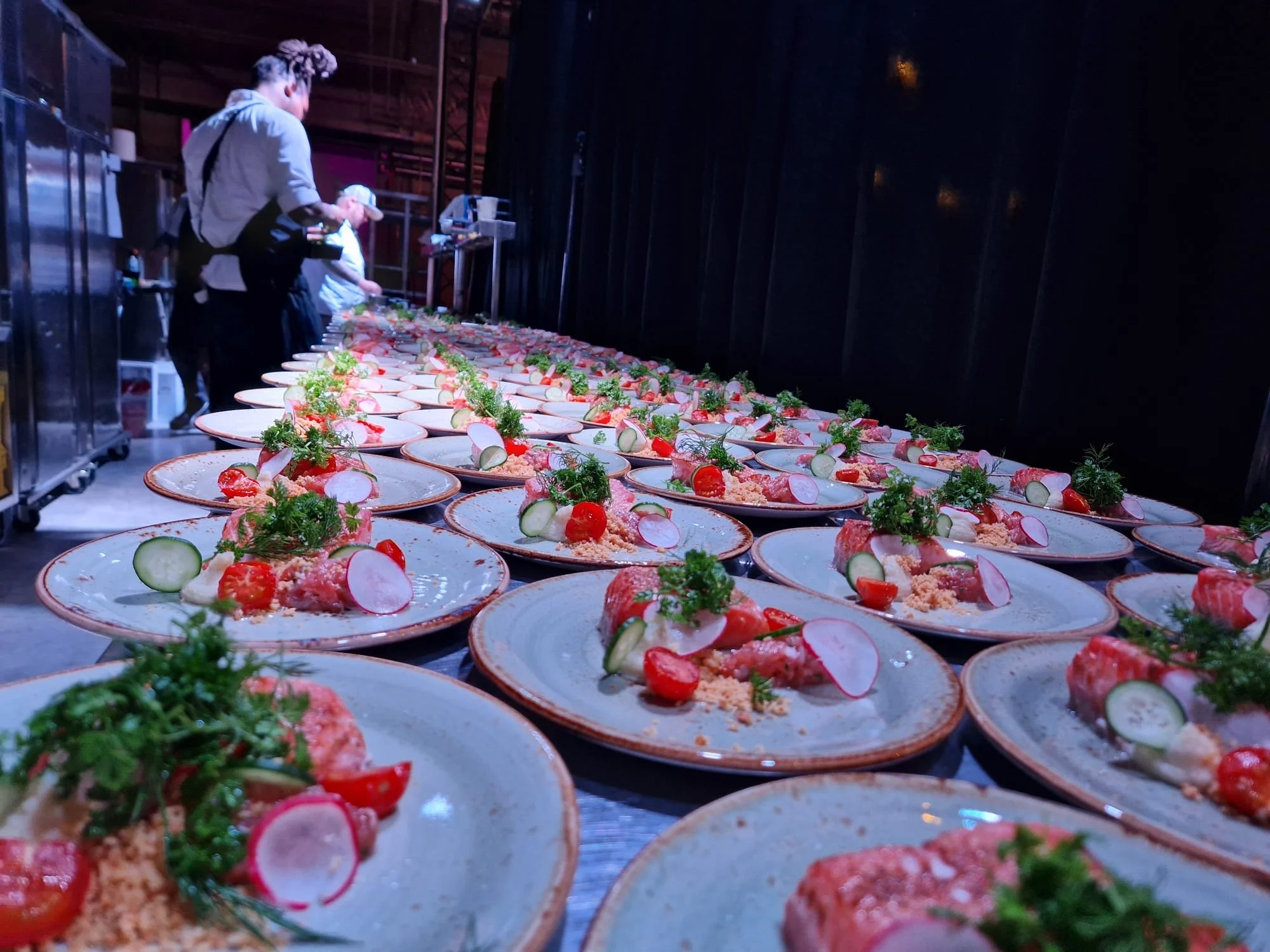 Multiple plates of food with sliced vegetables and garnishes, lined up on a long table at a catering event.