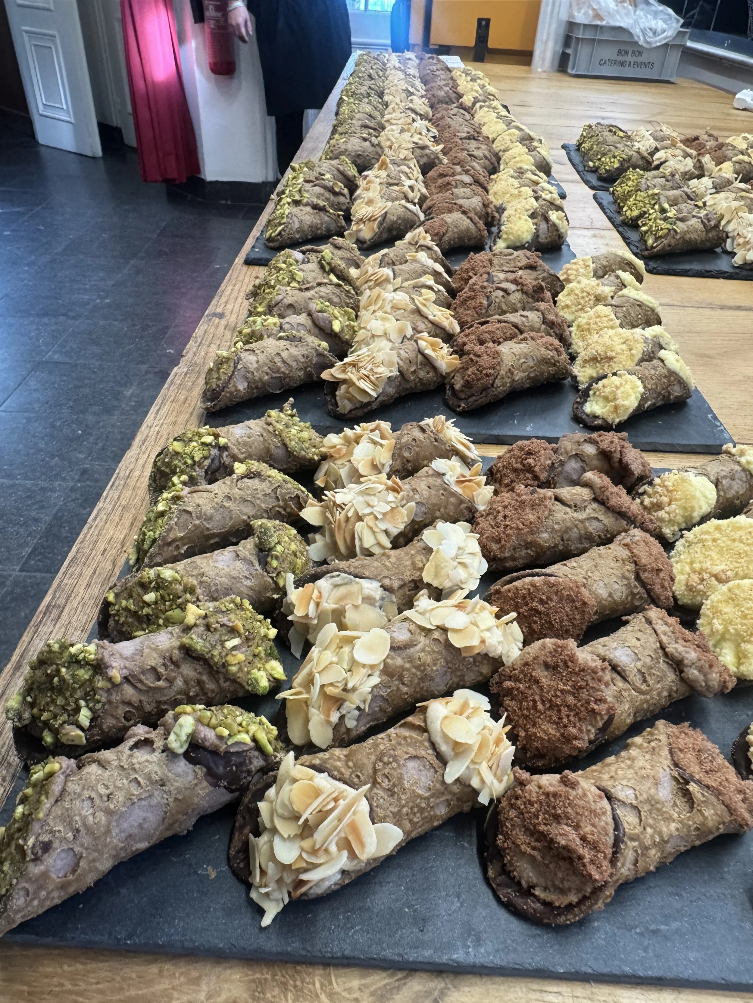 Assorted cannoli on a long black slate platter on a wooden table, decorated with chopped nuts and pistachios.