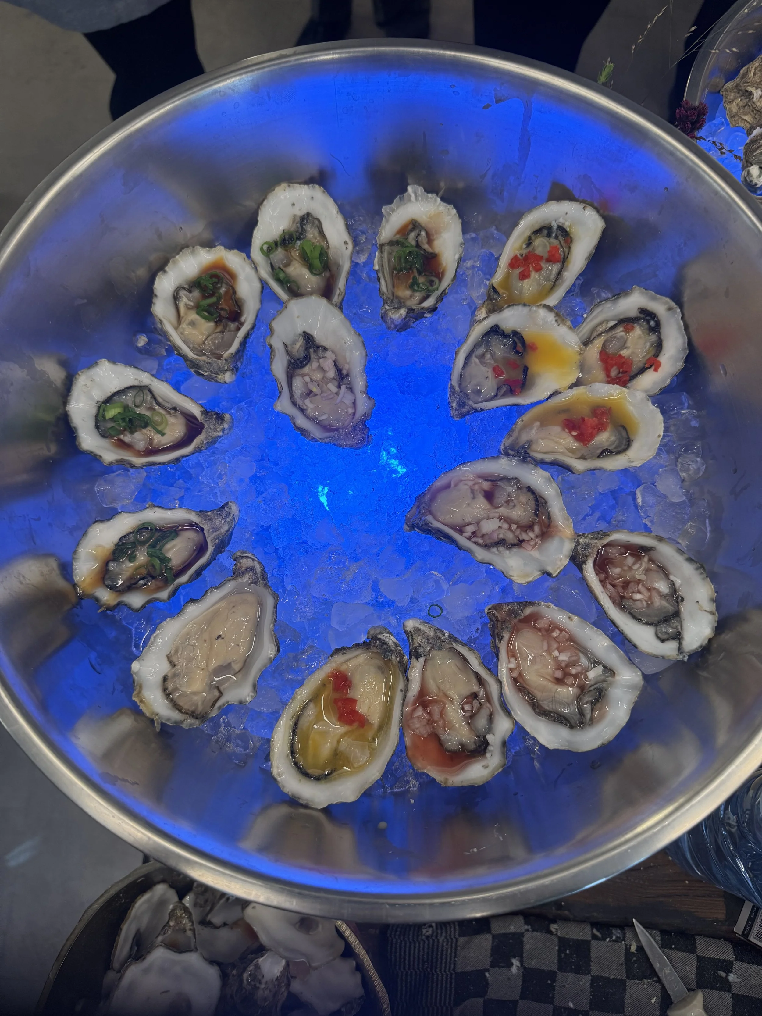 A round metal container filled with ice, holding open oyster shells topped with various garnishes such as chopped green onions and red peppers, illuminated by blue light.